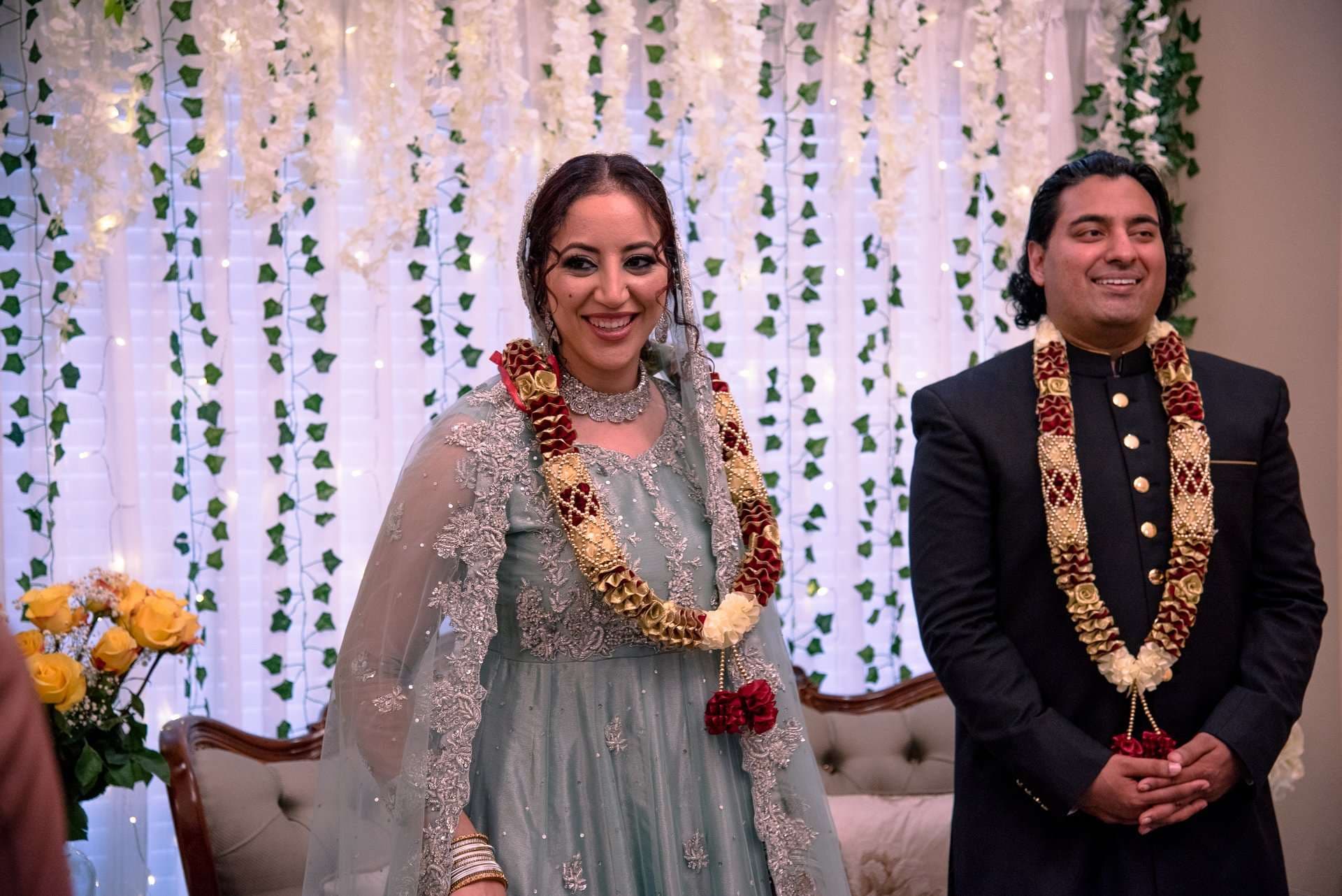 Smiling couple wearing floral garlands, she in a sage green embroidered gown and he in a black jacket, before an ivy backdrop