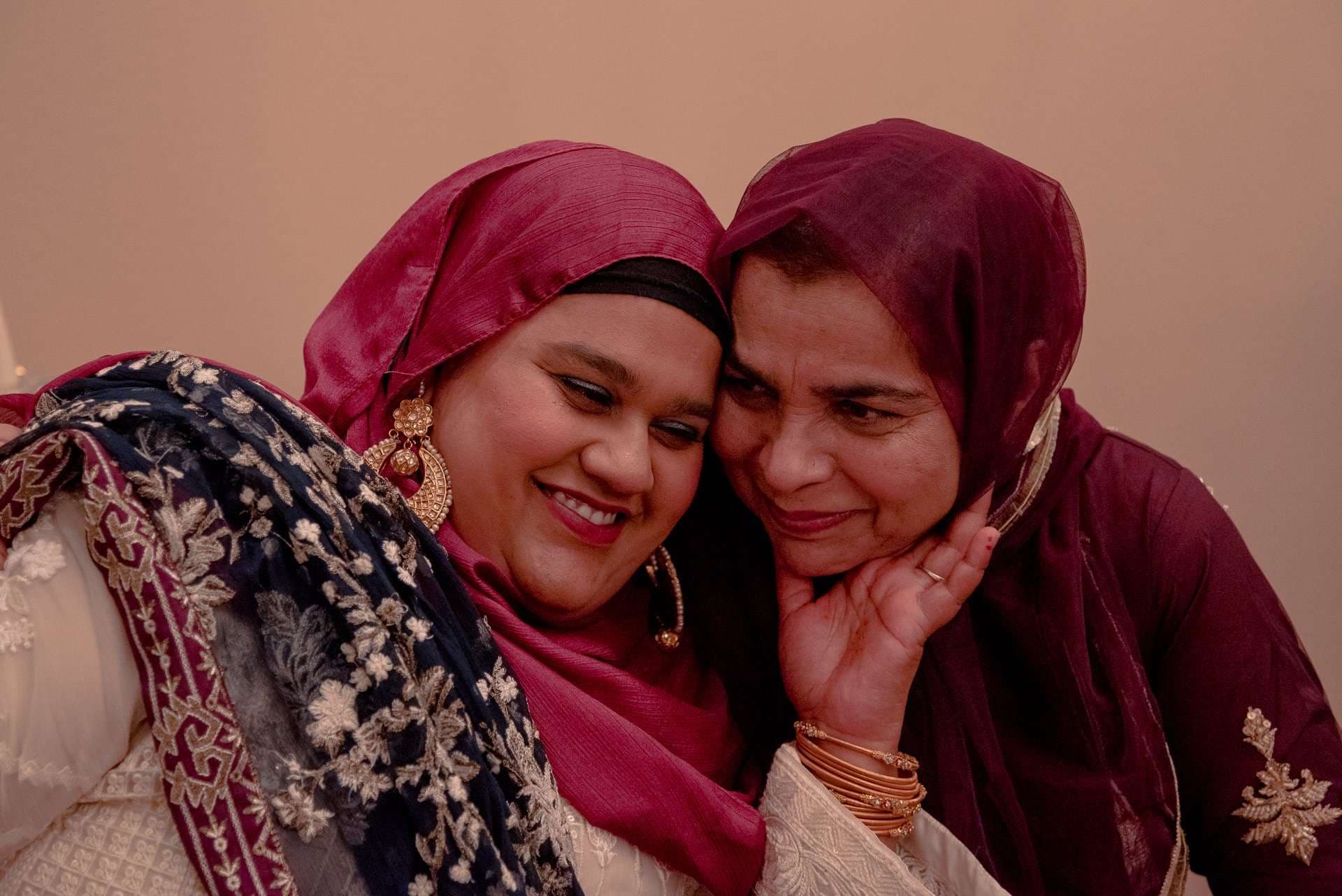 Two smiling women in magenta and maroon headscarves with embroidered shawls leaning their heads together affectionately