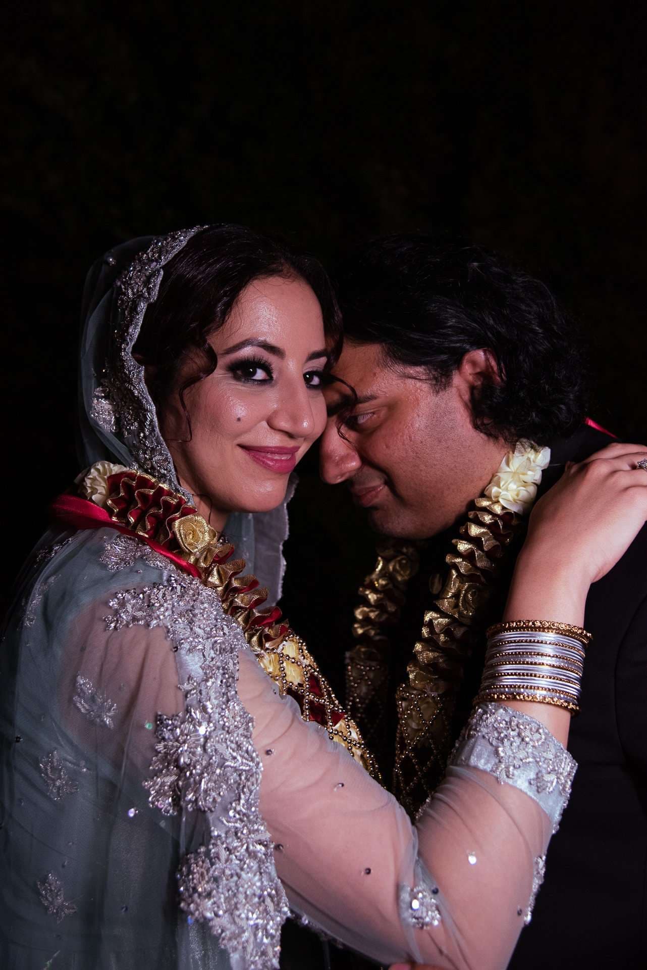 Couple pressing foreheads together in floral garlands, she in a embroidered gray-blue veil and sheer sleeves, wearing bangles