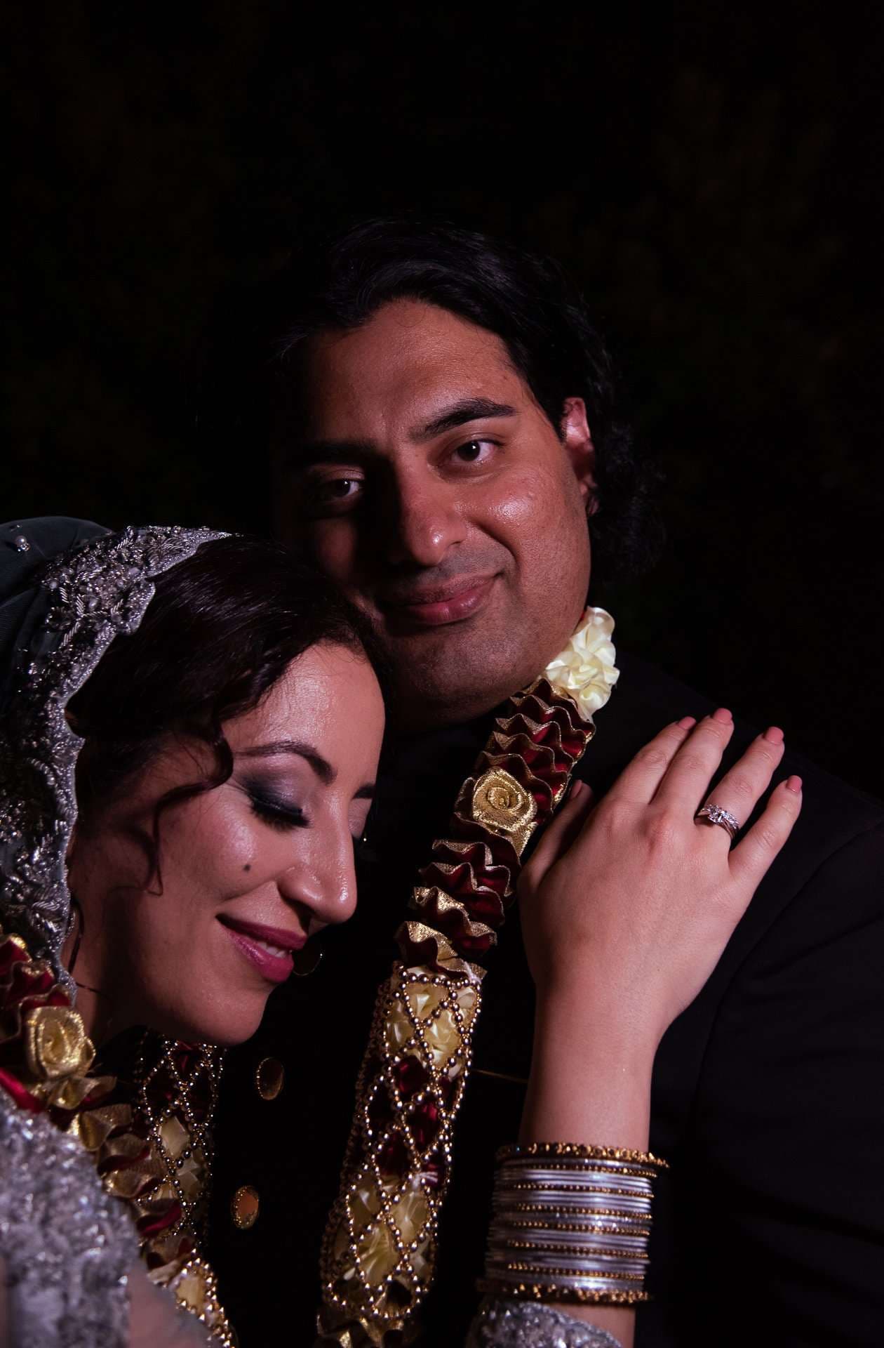 Couple in traditional wedding attire embracing against a dark background, her head on his chest, both wearing ornate garlands