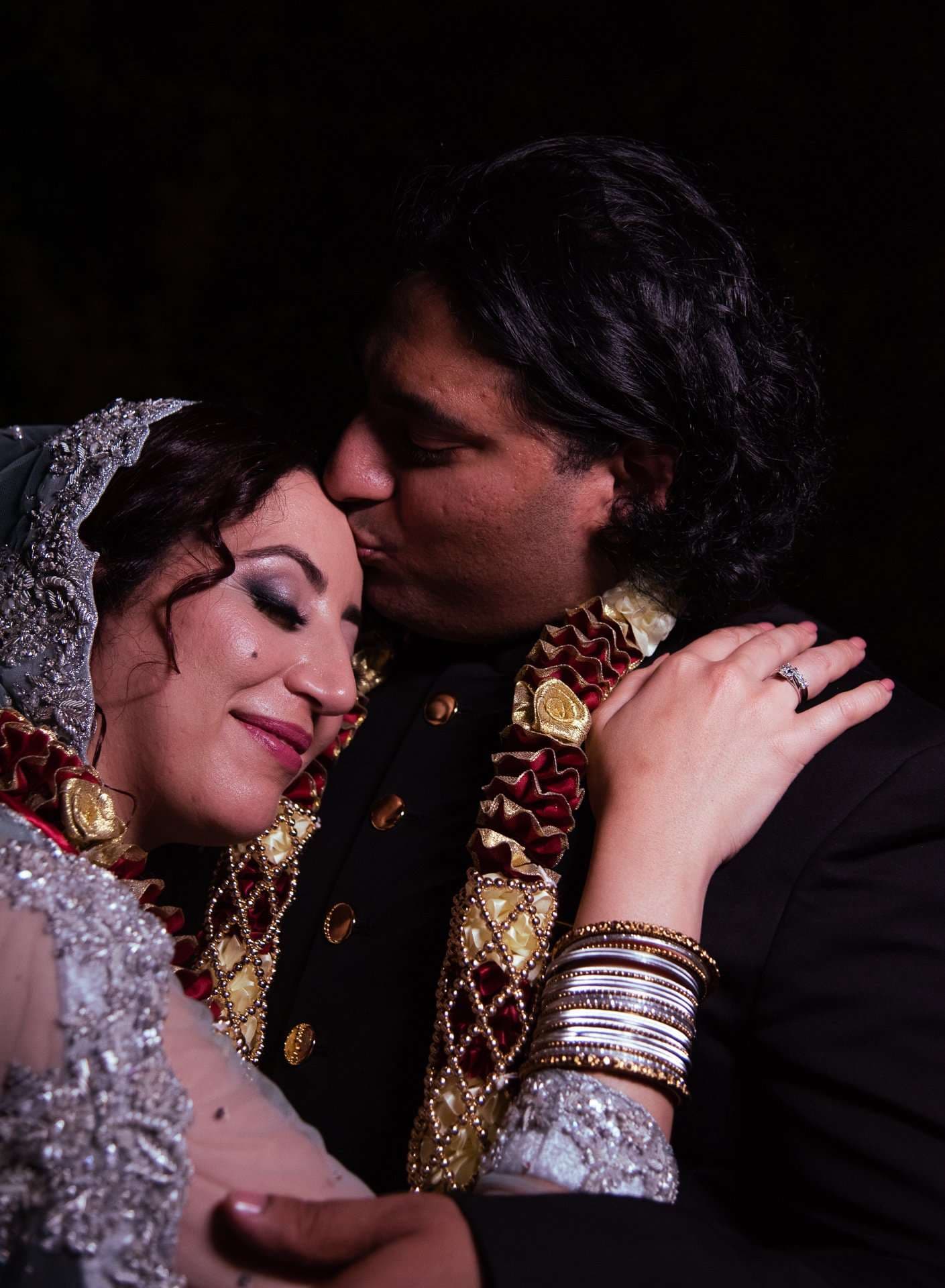 Groom kissing his smiling bride's forehead, both in ornate traditional wedding attire with red and gold garlands