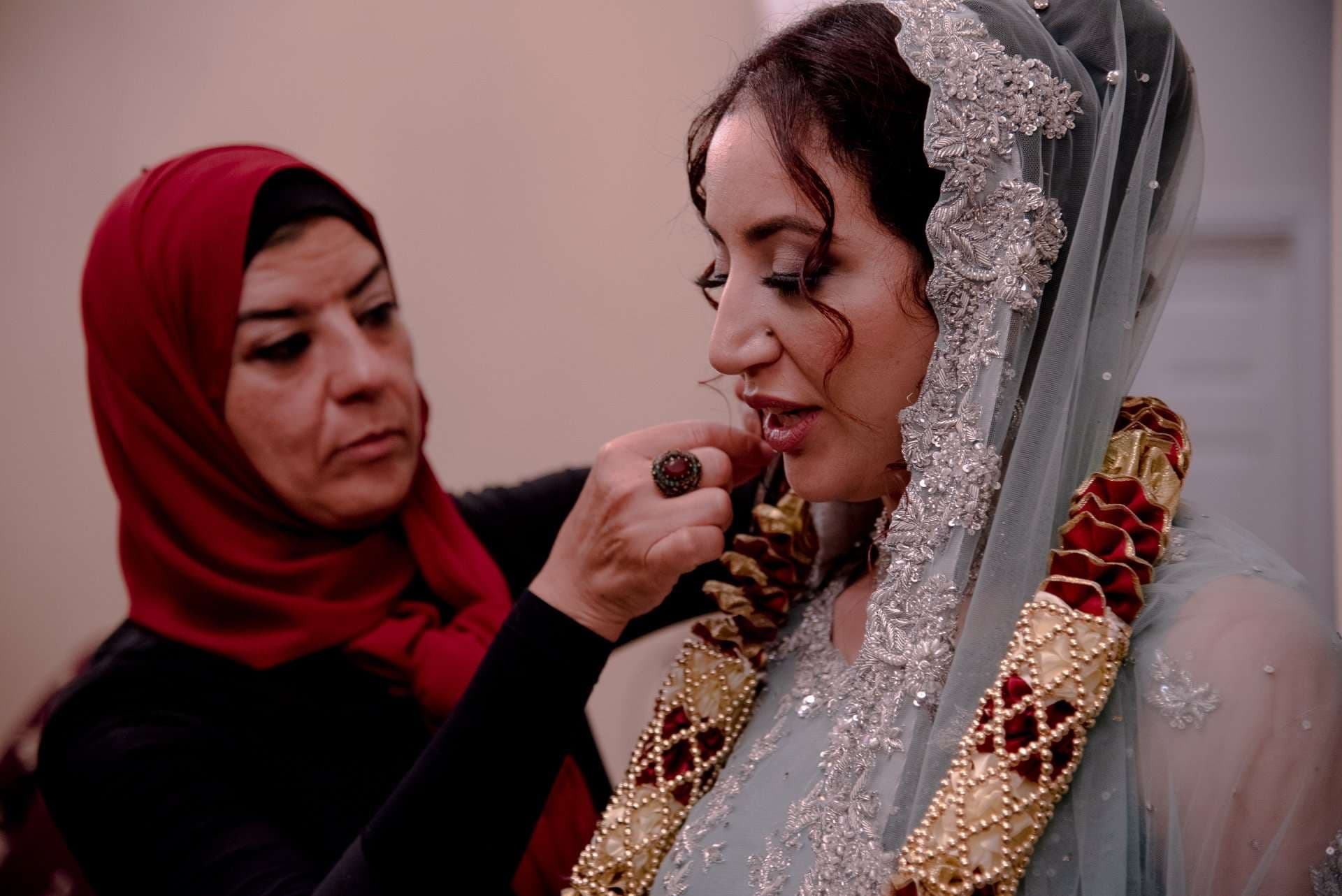 Woman in a red headscarf adjusts the jewelry of a bride in a lace-trimmed veil and pale blue gown