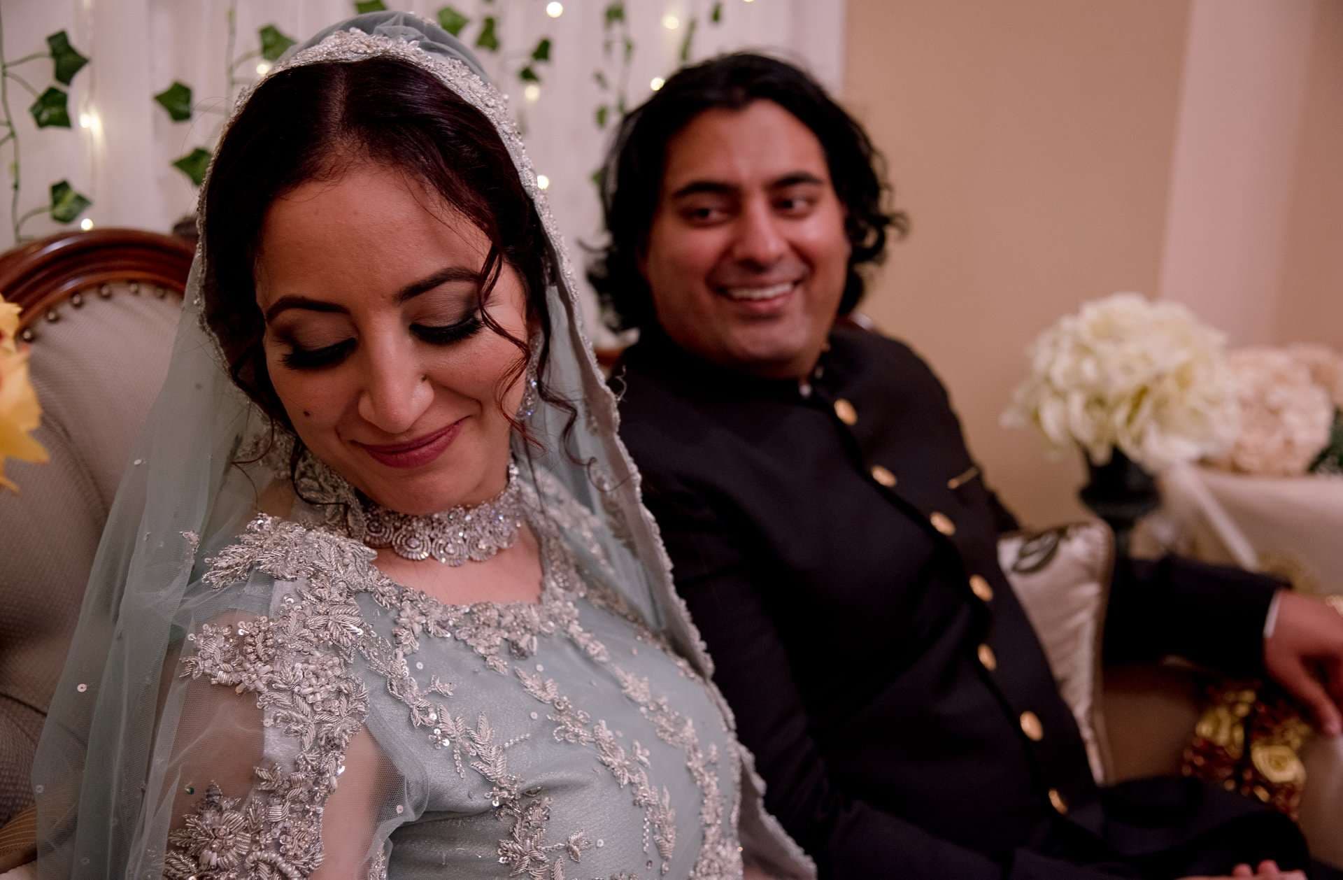 Smiling bride in a embroidered sage gown and veil looks down while a man in a black jacket beams behind her