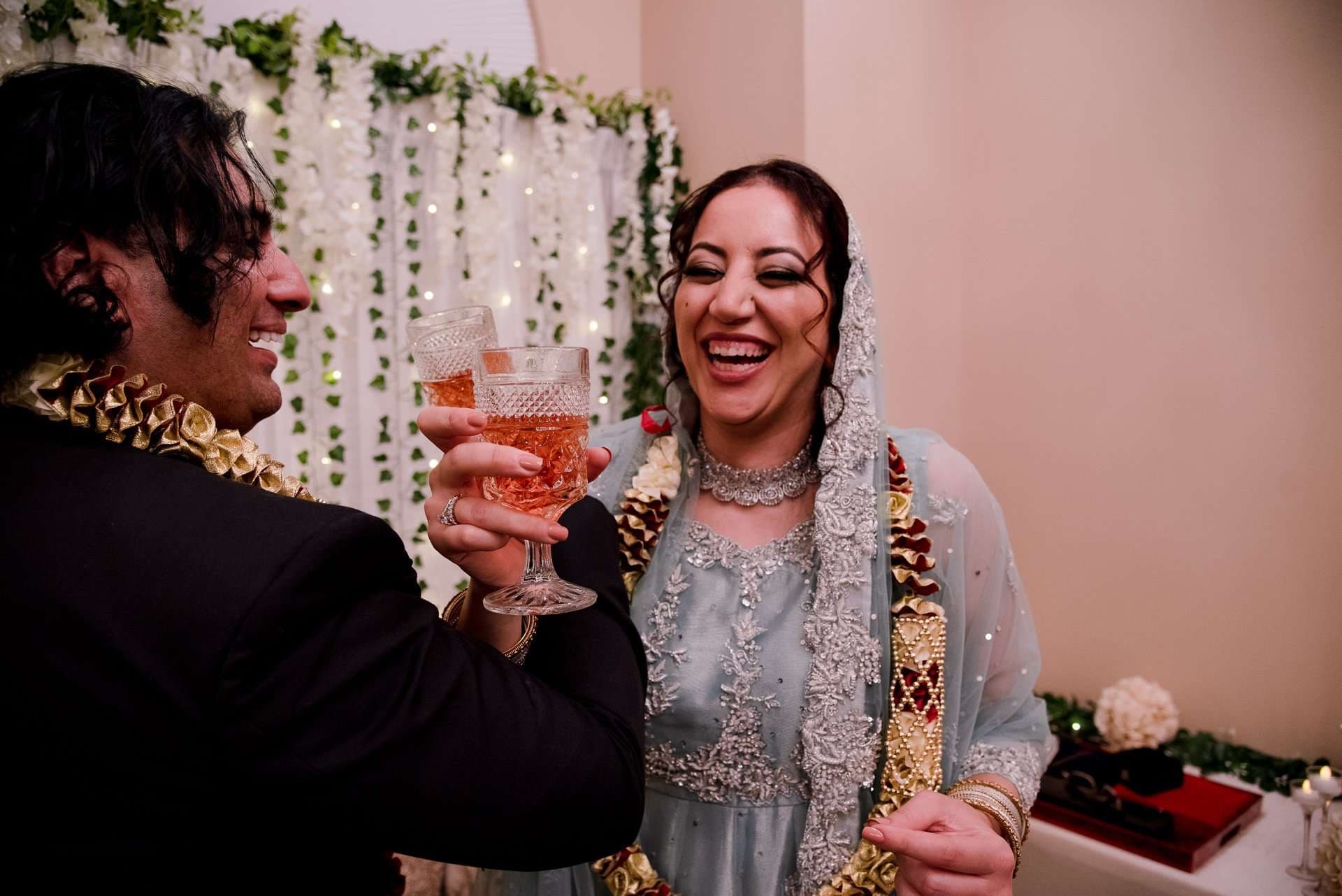 Laughing bride in a pale blue embroidered outfit and veil toasting with a man in a dark suit, both wearing garlands