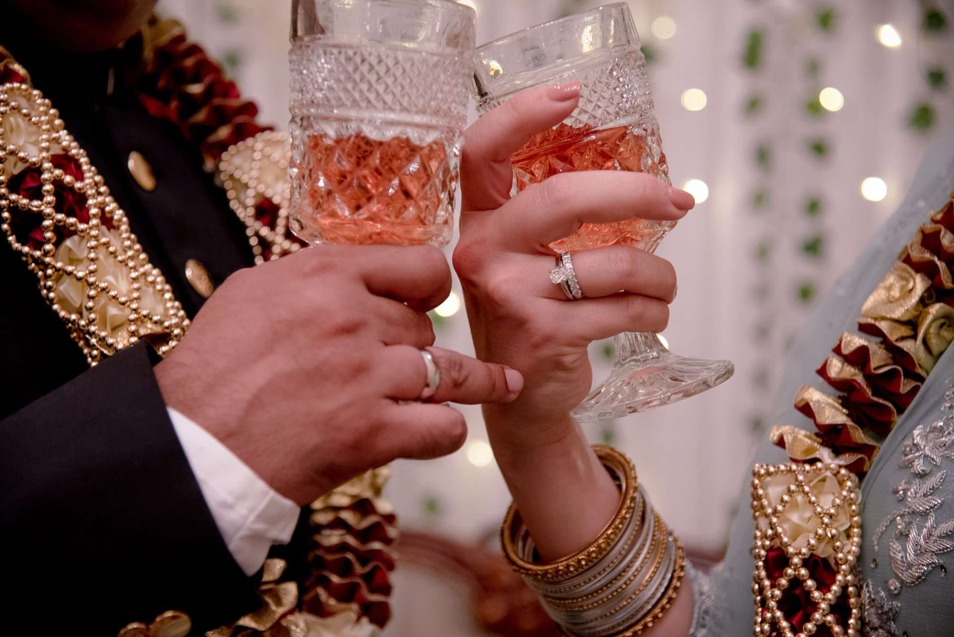 Couple clinking cut-crystal glasses of rosé, wearing rings, bangles and ornate embroidered wedding attire