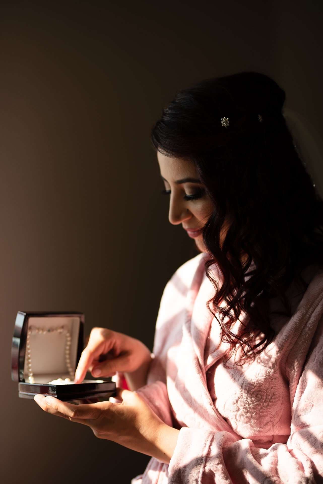 Woman in a pink robe with a flower hair clip smiles down at a pearl necklace in an open jewelry box