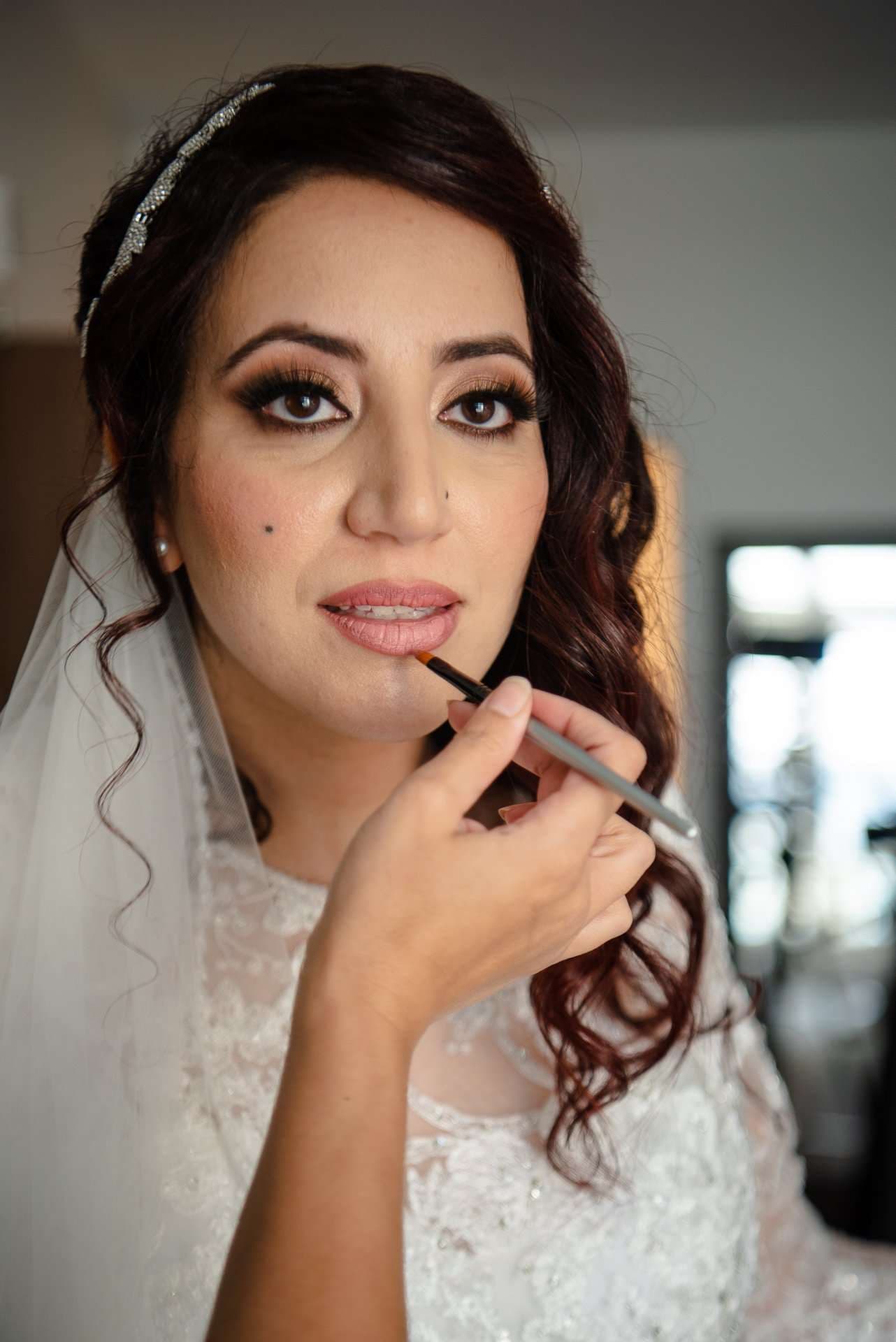 Bride in a lace gown and veil having her lip liner applied while getting ready for her wedding