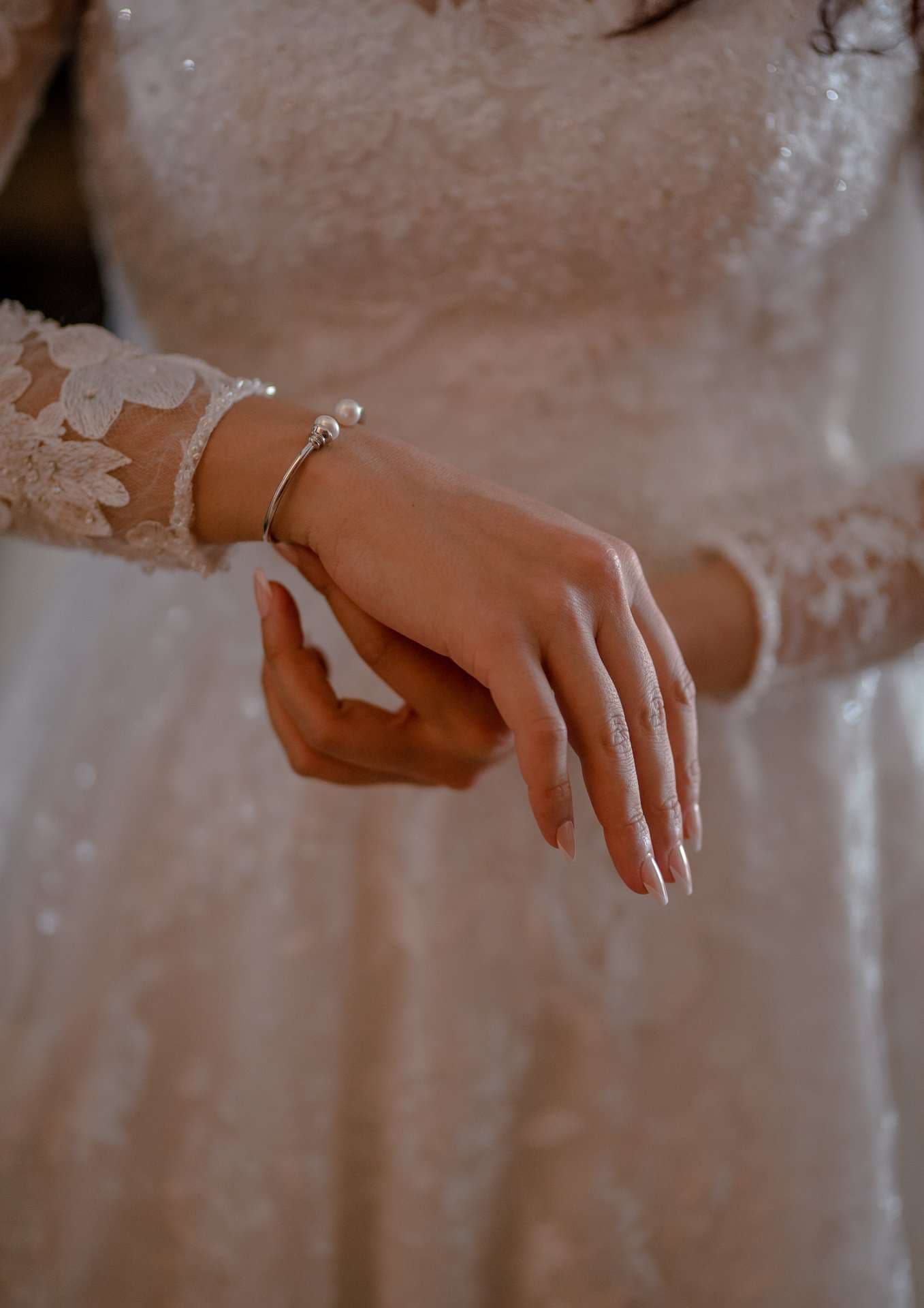 Bride in a long-sleeved white lace gown adjusting a silver pearl bracelet on her wrist