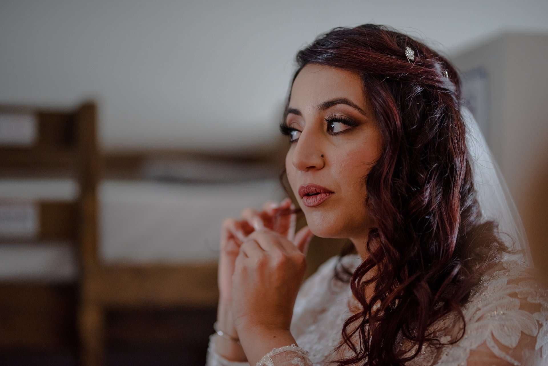 Bride in a lace gown and veil putting on an earring while gazing aside in a rustic room