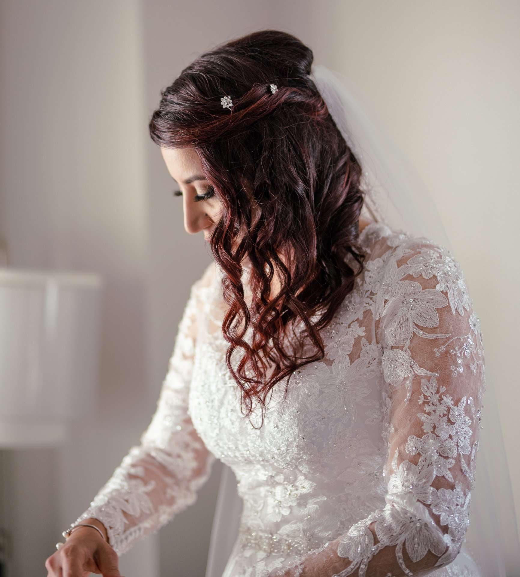 Bride in a long-sleeved white lace gown and veil looking down, her dark auburn curls pinned with small crystal accents