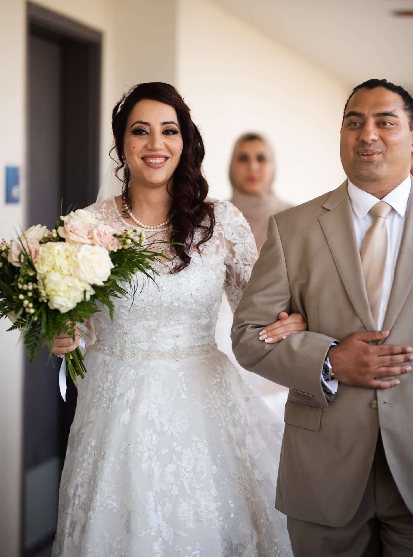 Smiling bride in a white lace gown holding a cream and pink bouquet, walking arm in arm with a man in a tan suit