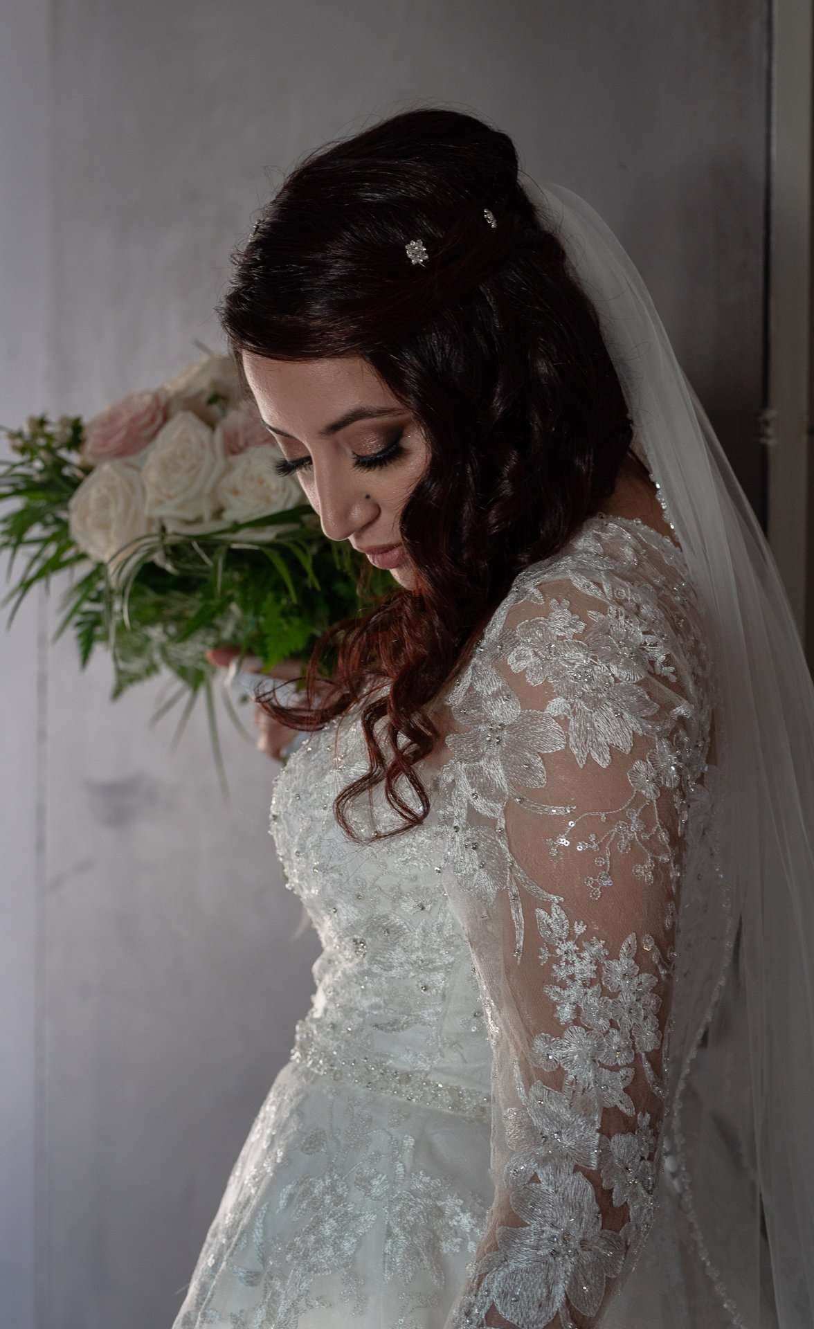 Bride in a white lace gown and veil looking down, holding a bouquet of white and blush roses