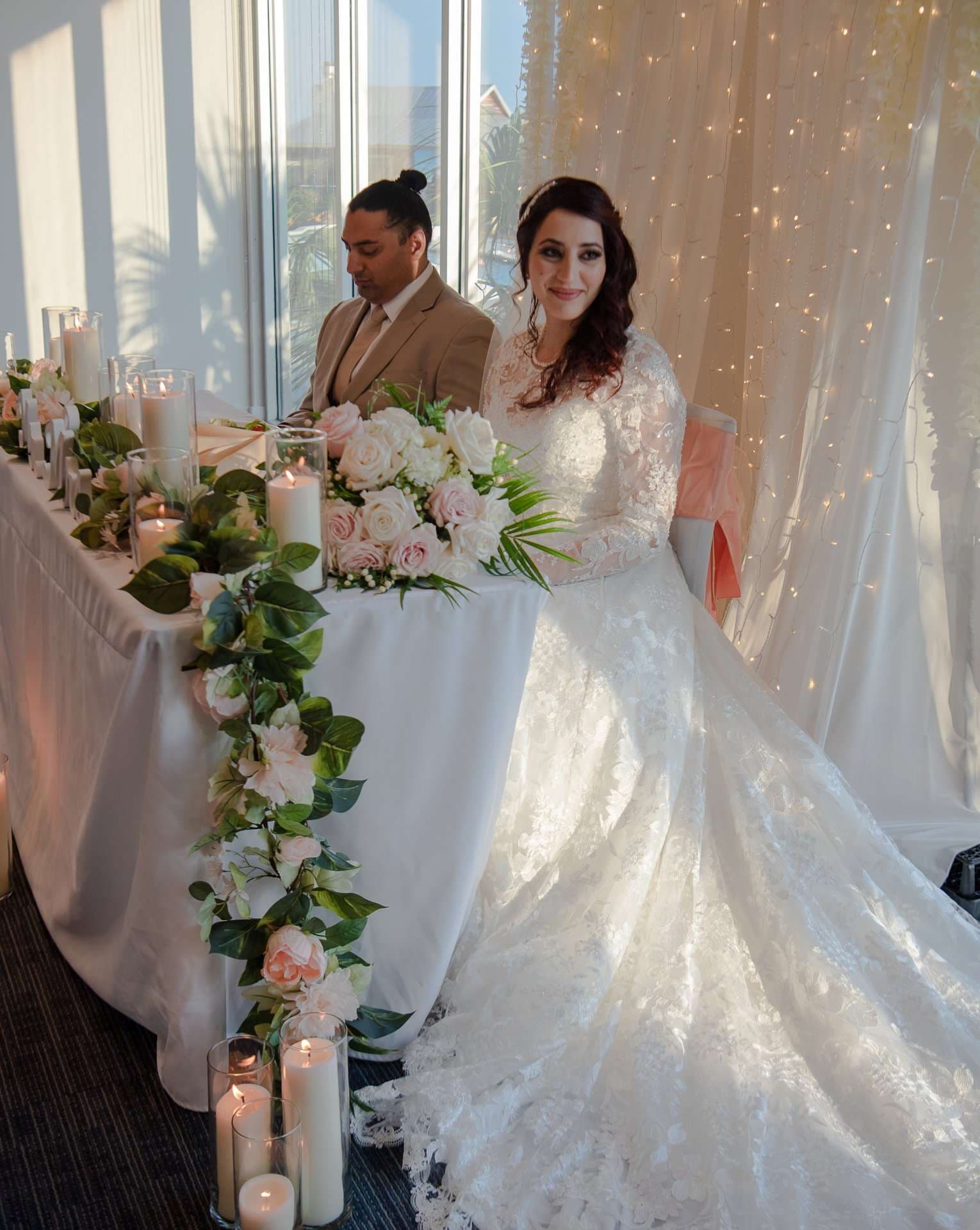 Couple seated at a sweetheart table with pink roses, greenery and candles, the bride in a lace gown beside fairy lights