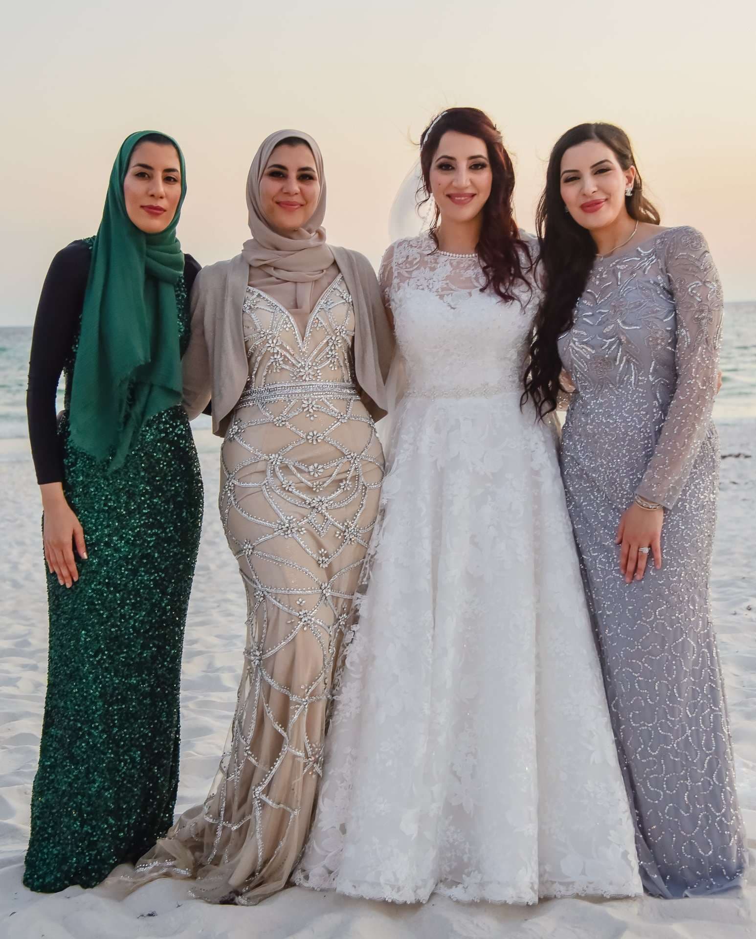 Bride in a white lace gown standing on the beach at sunset with three women in green, champagne and silver dresses