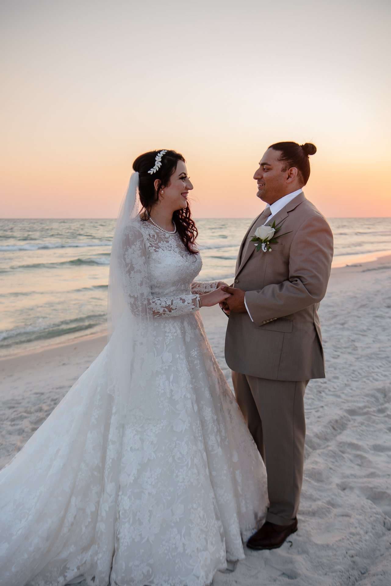Couple holding hands and smiling at each other on the beach at sunset, bride in a lace gown and veil