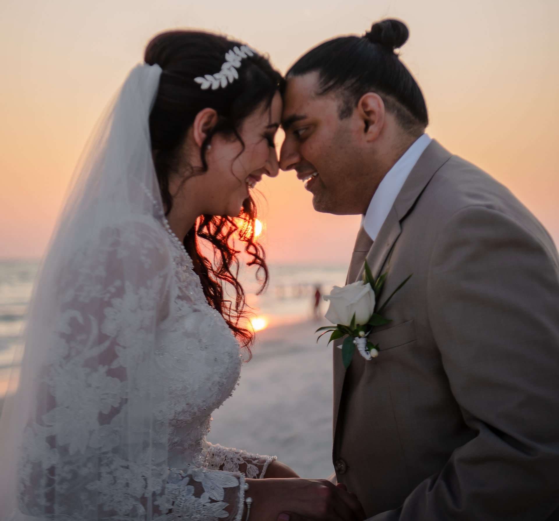 Bride in a lace gown and veil touching foreheads with her smiling groom on the beach at sunset
