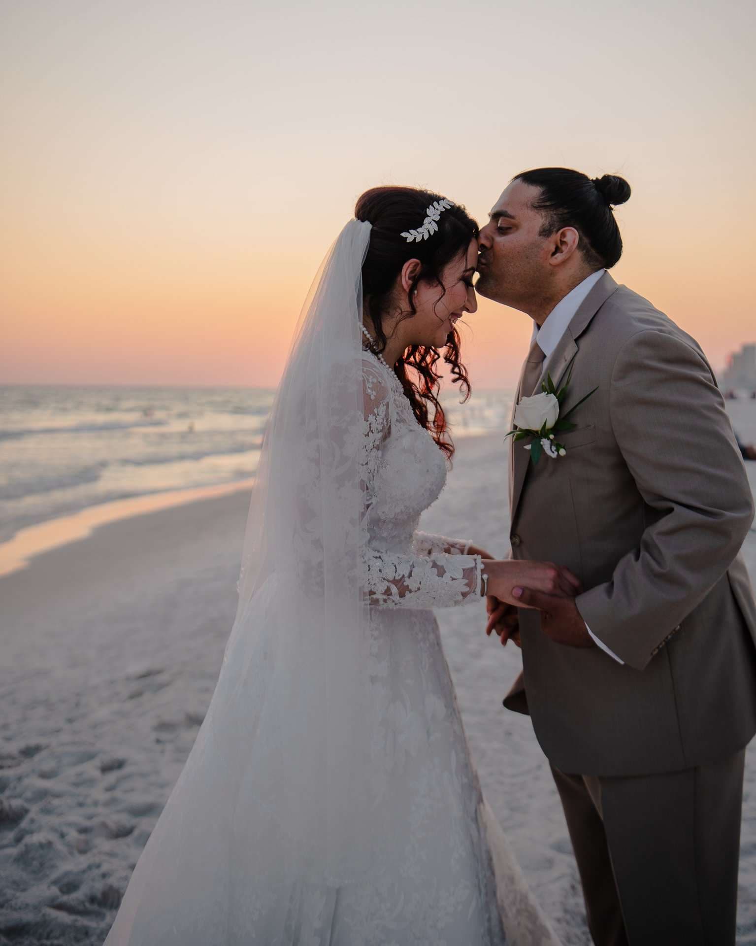 Groom kissing his bride's forehead on the beach at sunset, she wears a lace gown, veil and leaf hairpiece