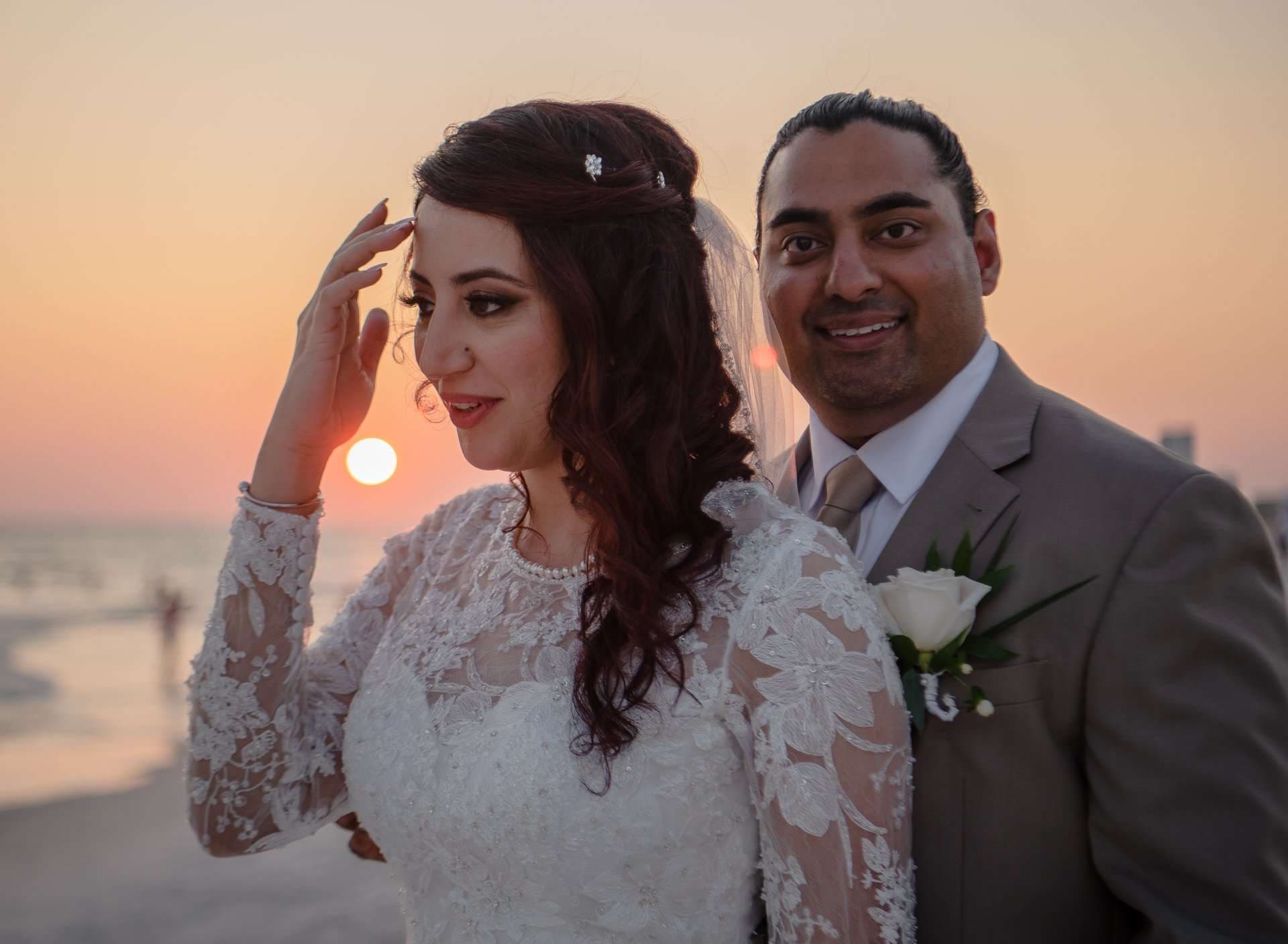 Bride in a white lace gown touching her hair while her smiling groom stands behind her at sunset on the beach