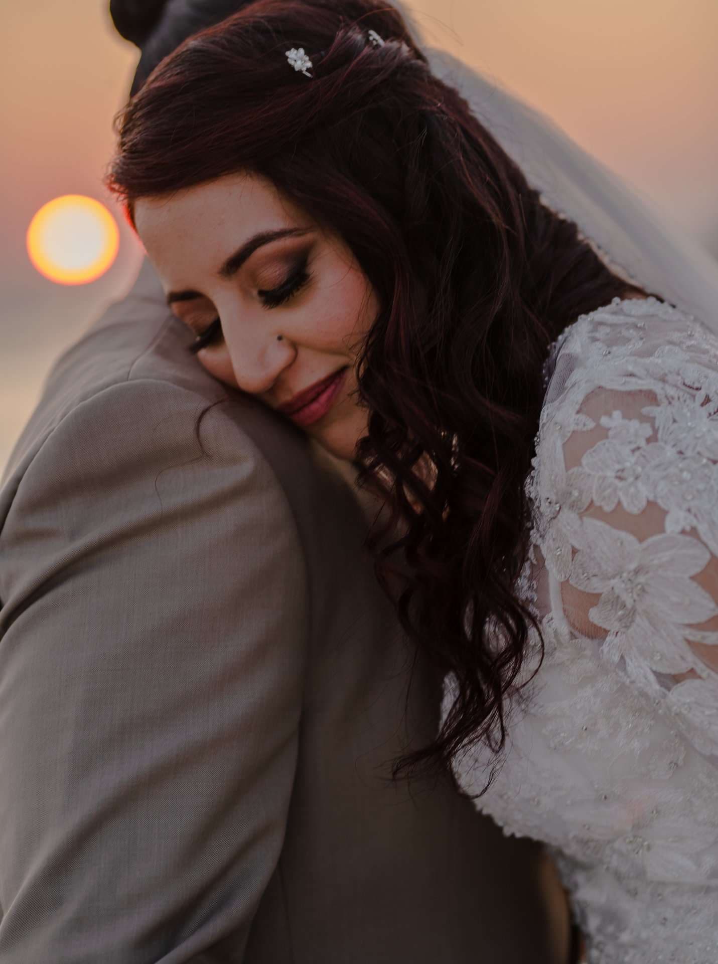 Bride in a lace gown and veil resting her head on her groom's shoulder as the sun sets behind them