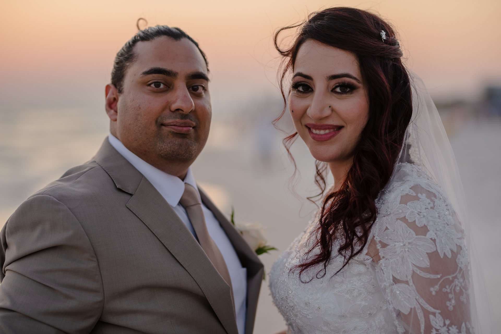 Couple in a tan suit and lace wedding gown with veil smiling toward the camera on a beach at sunset