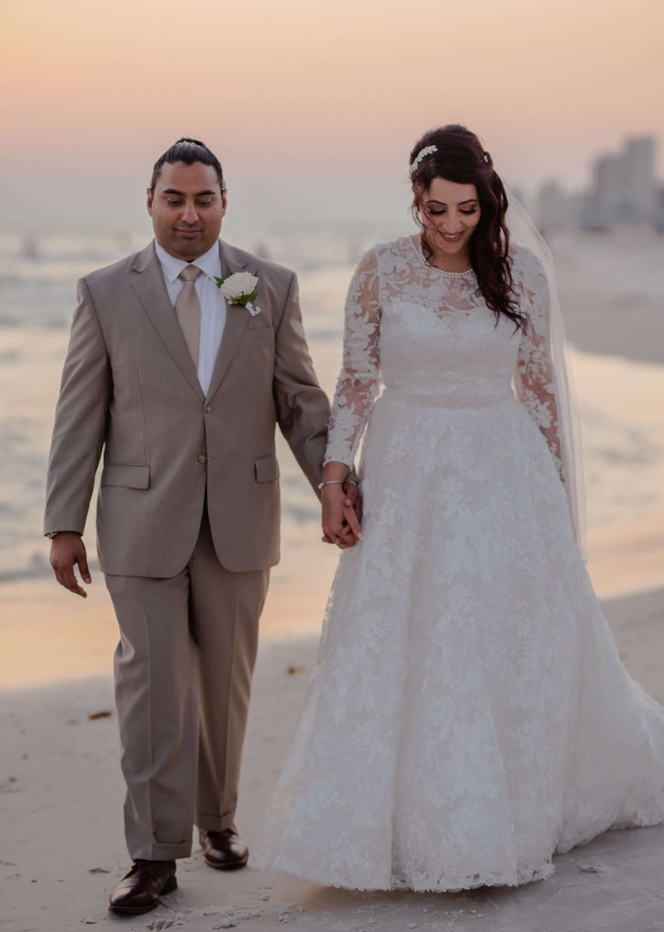 Couple holding hands walking along the beach at sunset, groom in a tan suit and bride in a lace gown with veil