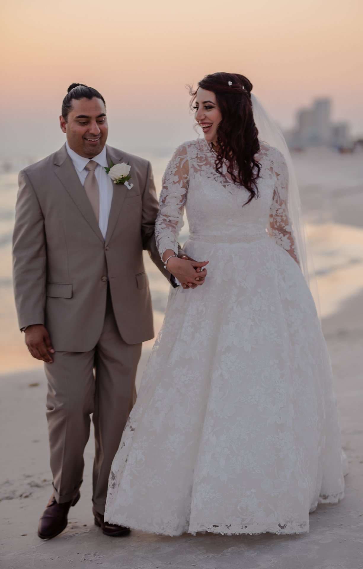 Couple in a tan suit and white lace gown walking hand in hand on the beach at sunset, smiling