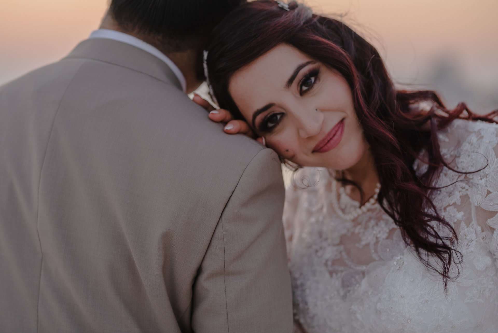 Bride in a white lace gown smiling and resting her head on her partner's shoulder at sunset