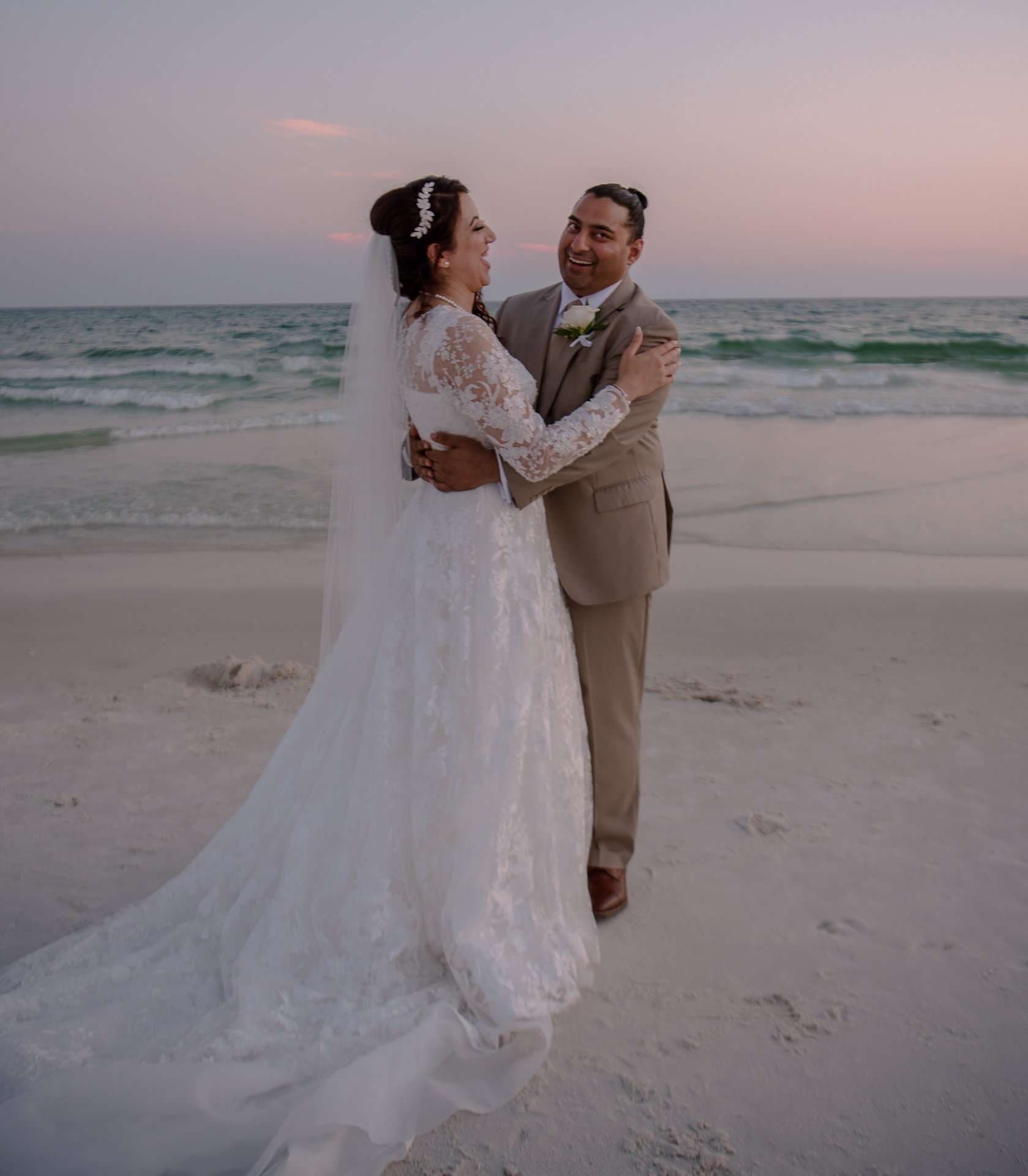 Bride in a lace gown and veil embracing her laughing groom in a tan suit on the beach at sunset