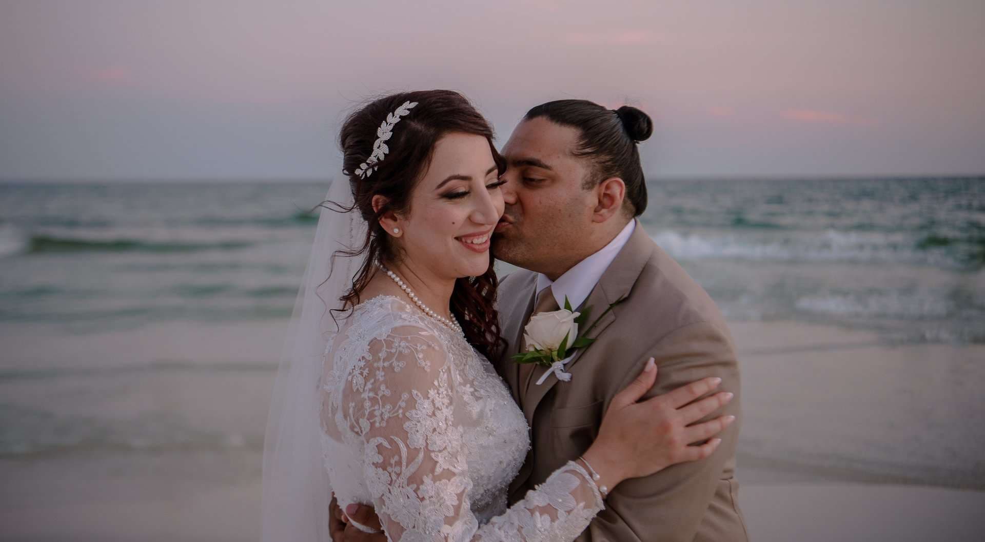 Groom in a tan suit kisses his smiling bride's cheek on the beach at dusk, she wears a lace gown and veil