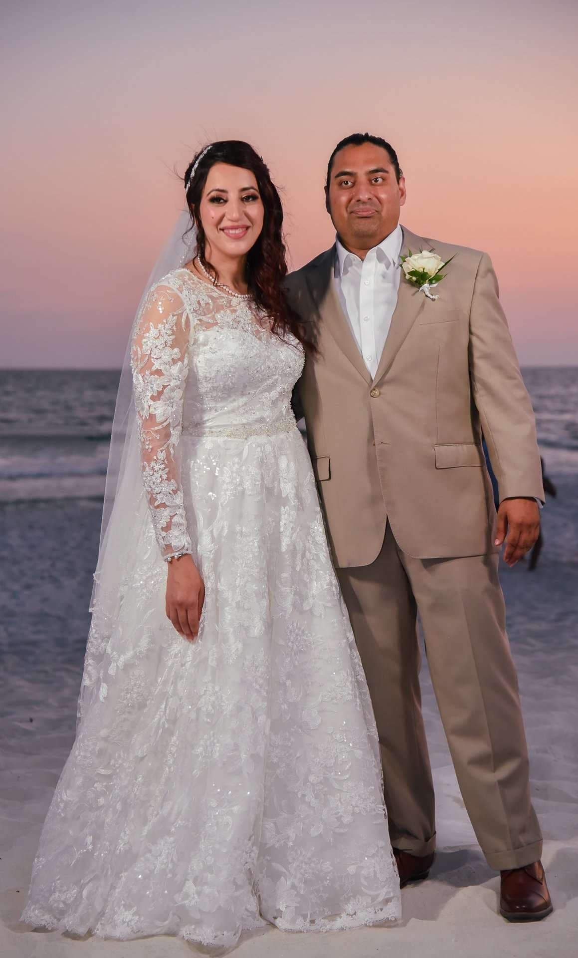 Couple posing on the beach at dusk, bride in a long-sleeved white lace gown and veil, groom in a tan suit
