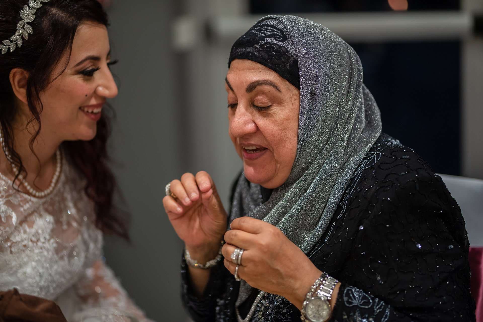 Smiling bride in a lace gown and beaded headpiece talks with an older woman in a shimmery gray hijab