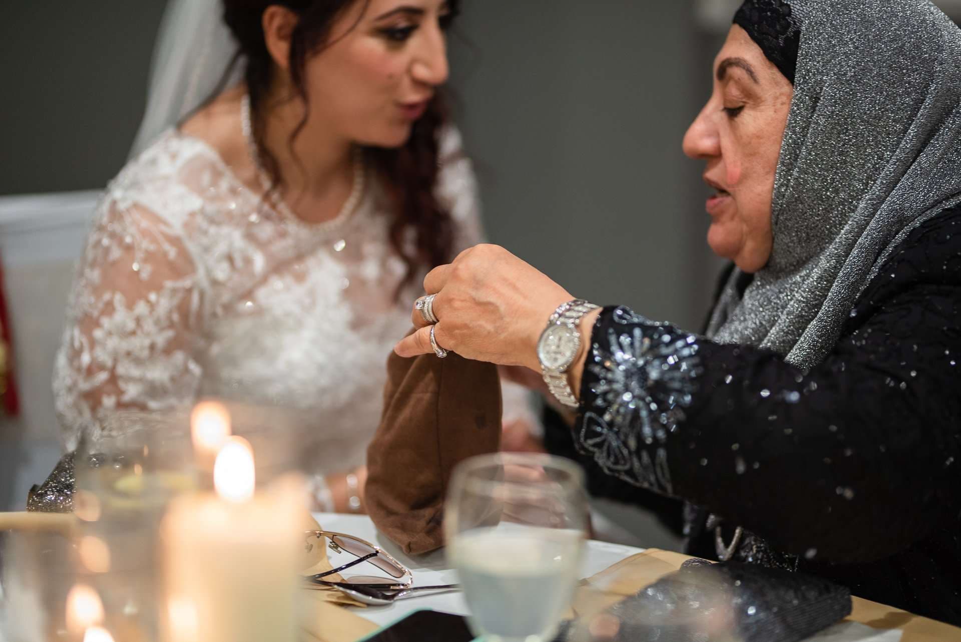 Bride in a white lace gown listens as an older woman in a silver headscarf holds a brown cloth pouch beside candles