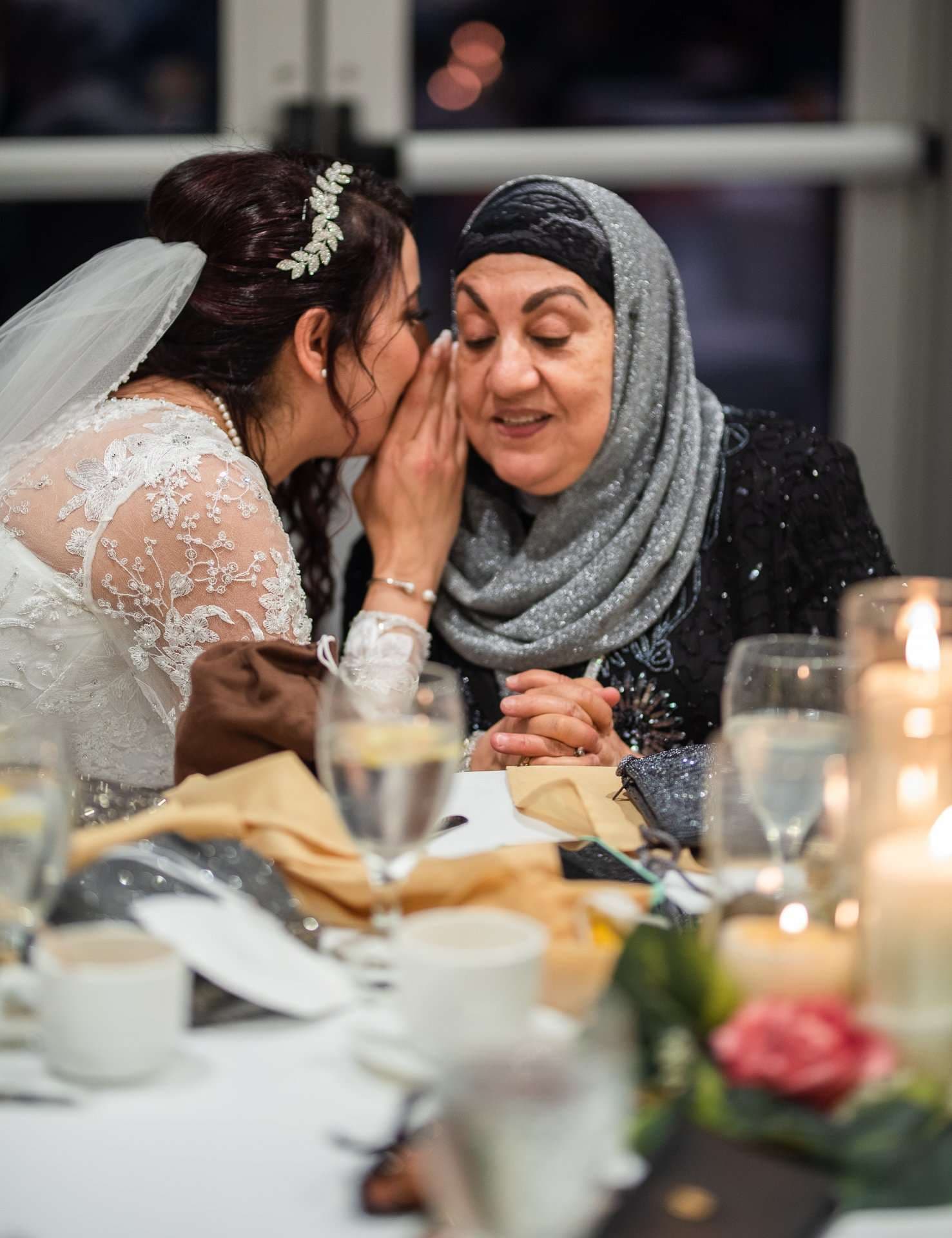 Bride in lace gown and veil whispering into the ear of a smiling older woman in a gray hijab at a reception table