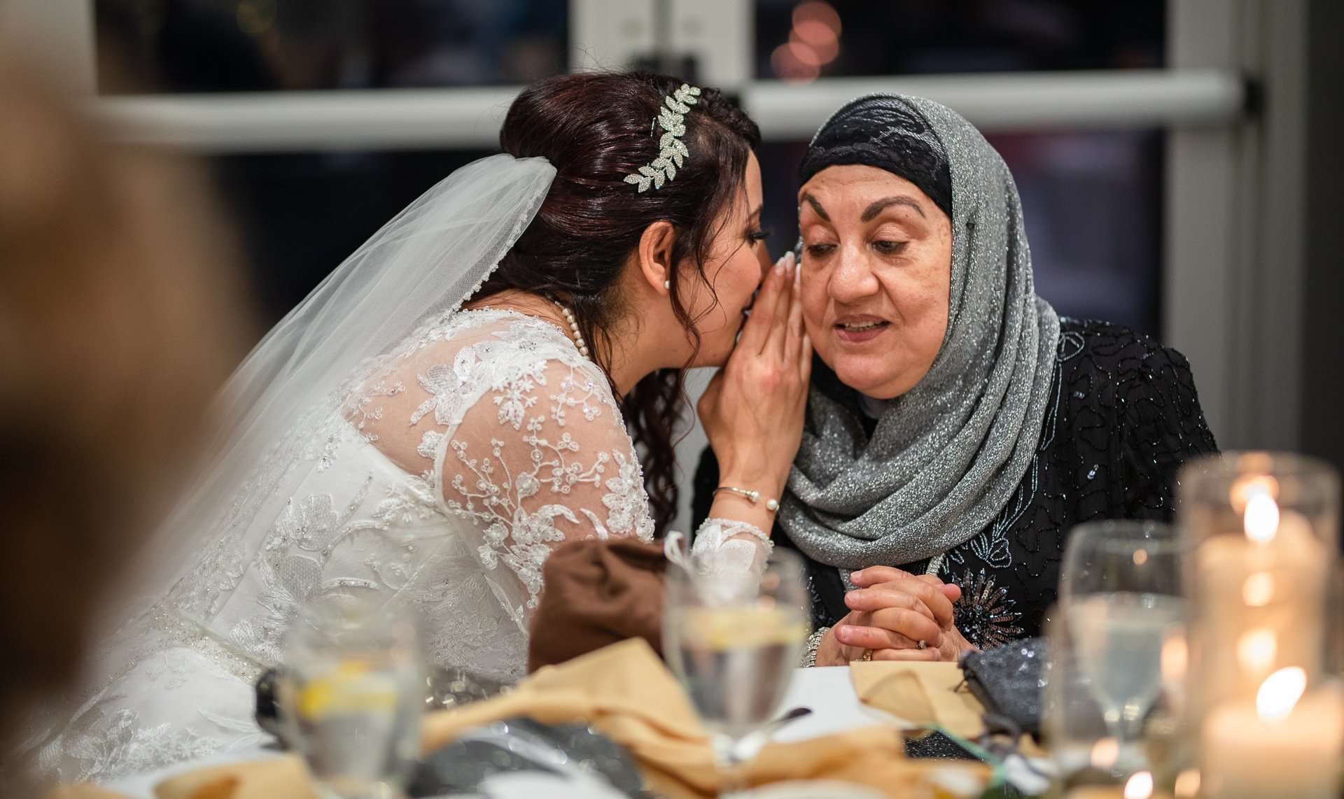 Bride in lace gown and veil whispers into the ear of an older woman in a gray hijab at a reception table
