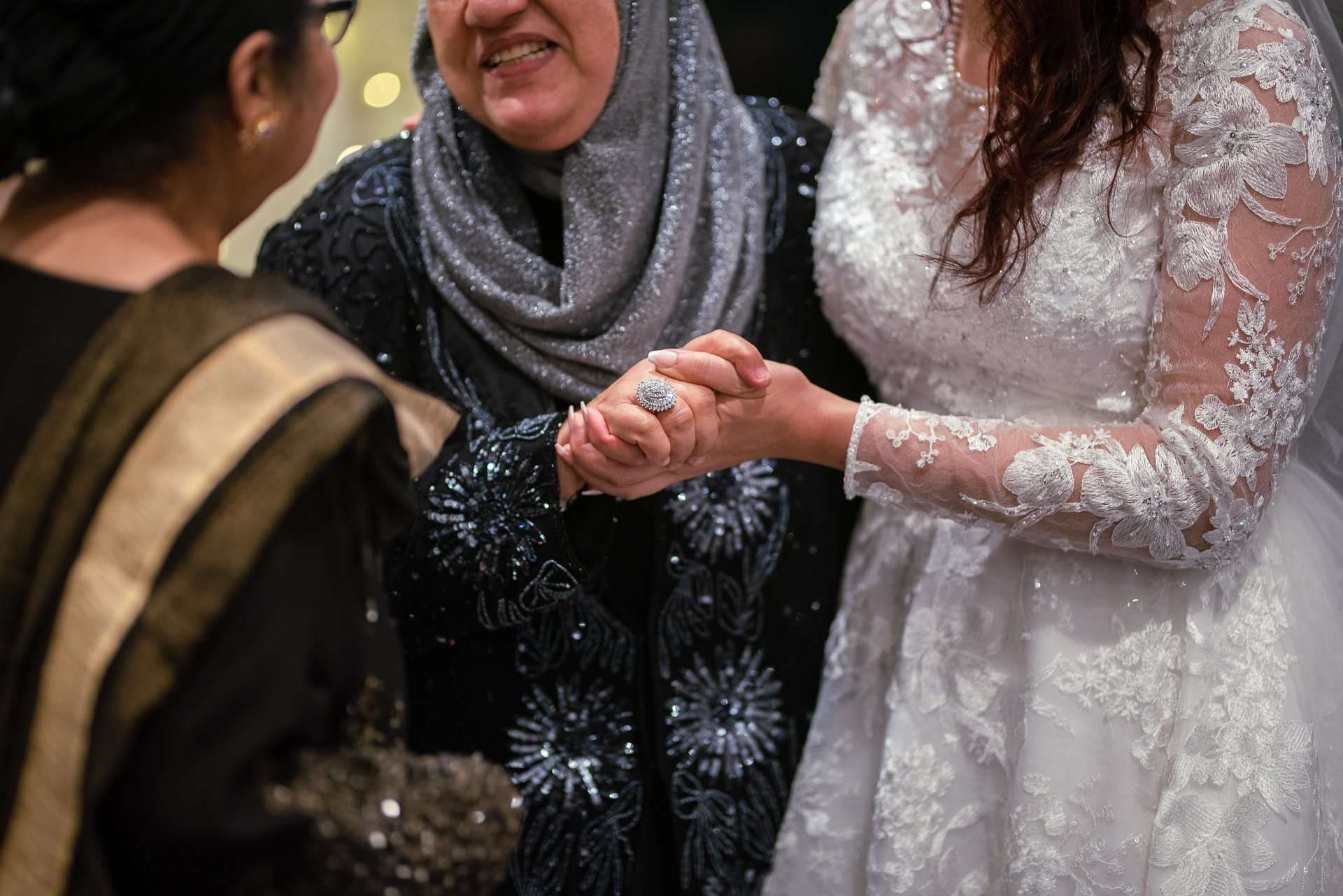 Bride in a lace gown clasping hands with a smiling woman in a gray sparkly headscarf and beaded dark jacket