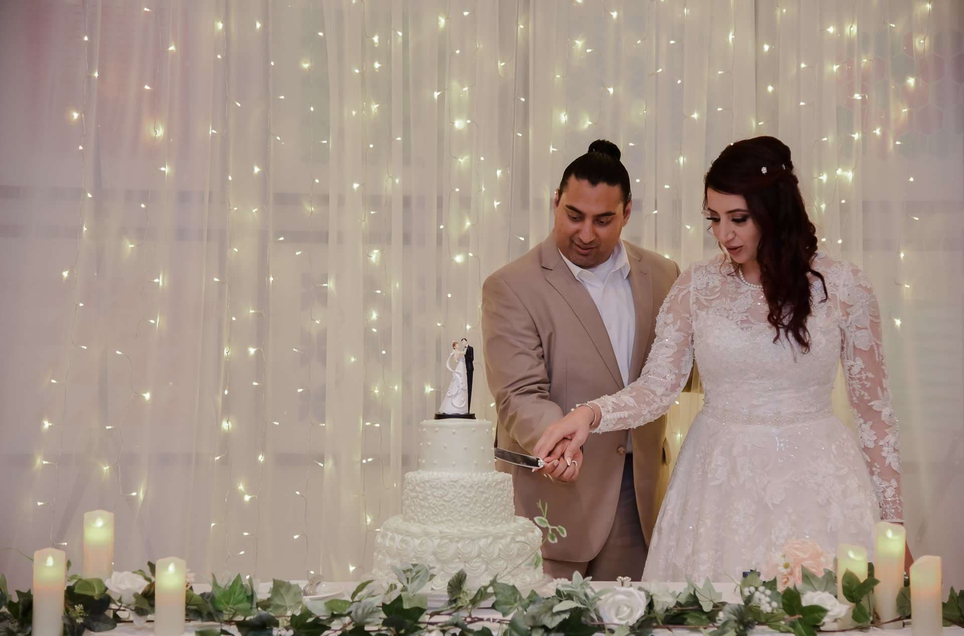 Couple cutting their three-tier white wedding cake together in front of a curtain of twinkling string lights