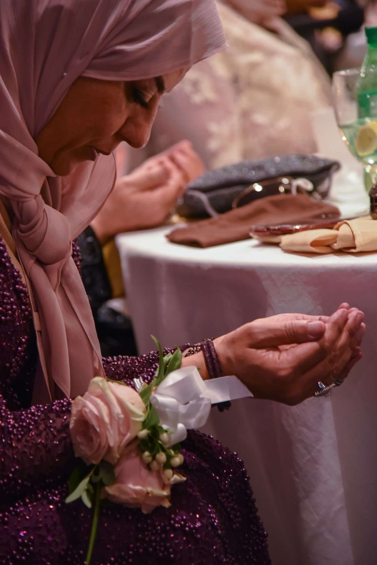 Woman in a blush hijab and beaded plum dress with a rose wrist corsage holds her hands open in prayer at a reception