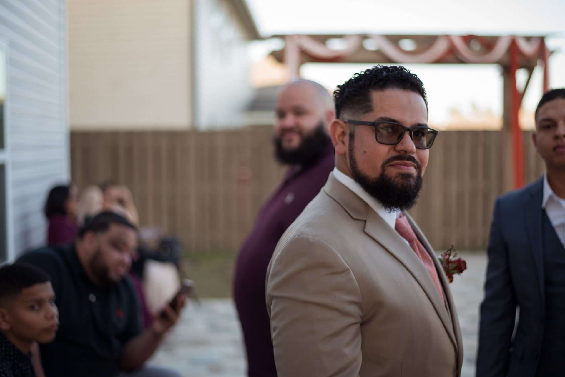 Bearded man in tinted glasses, tan suit and pink tie glances back at an outdoor wedding gathering