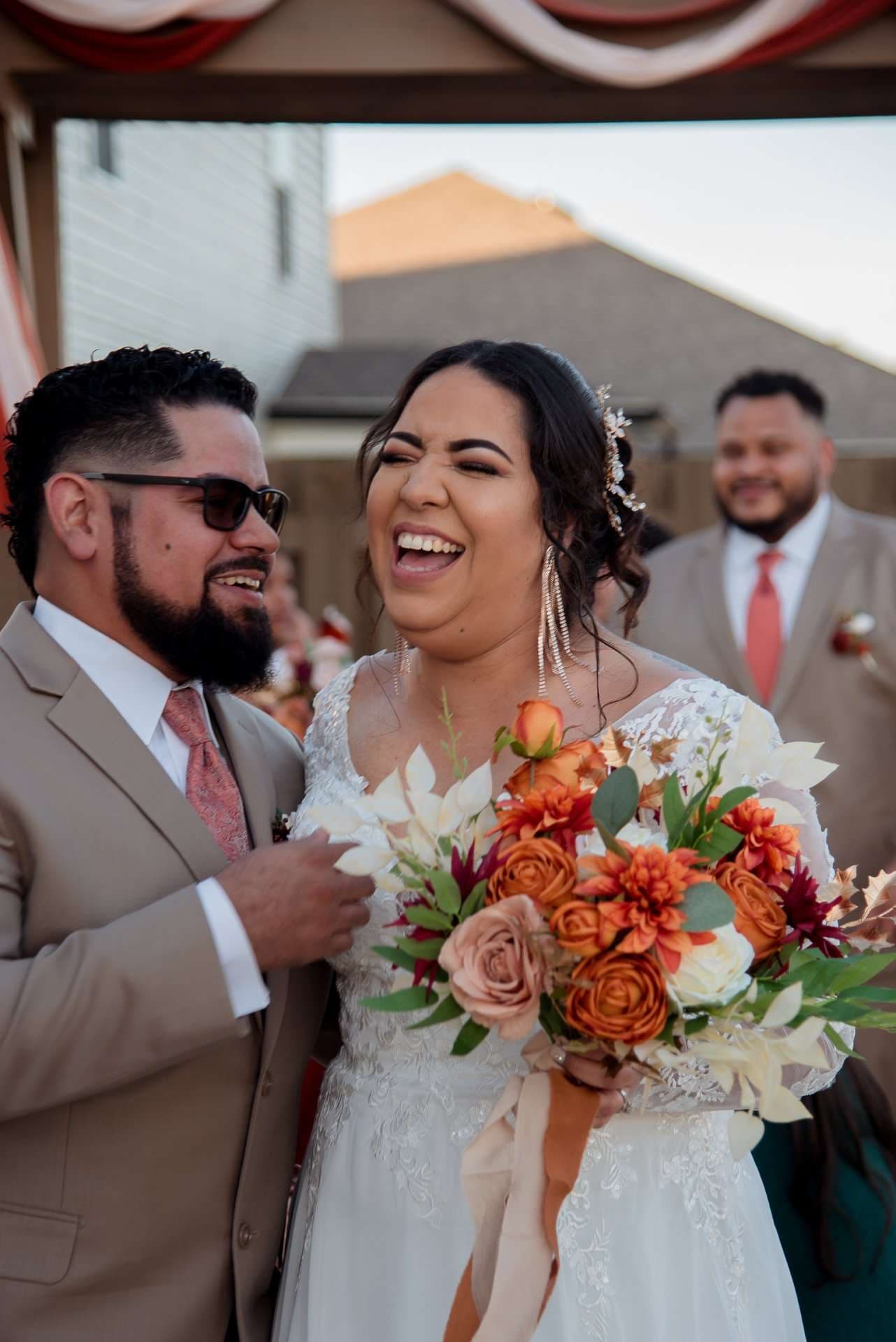 Laughing bride in a lace gown holding an orange and white bouquet beside a bearded groom in a tan suit