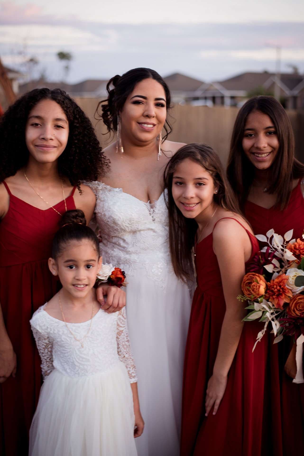 Bride in a white lace gown posing with three girls in red dresses and a flower girl, holding an orange bouquet