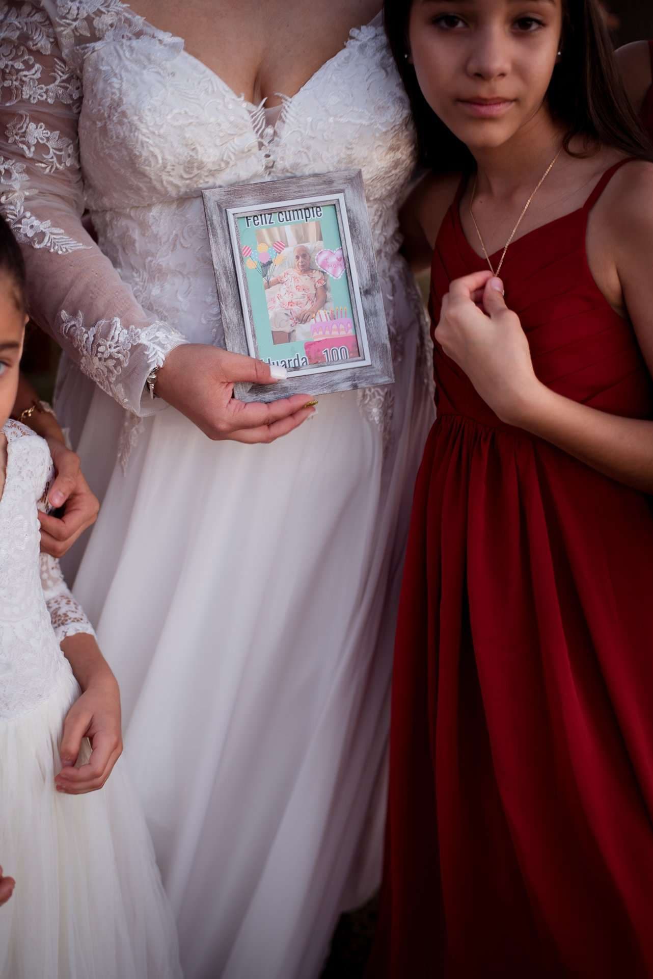Bride in a lace gown holding a framed birthday card, standing between two girls in white and red dresses
