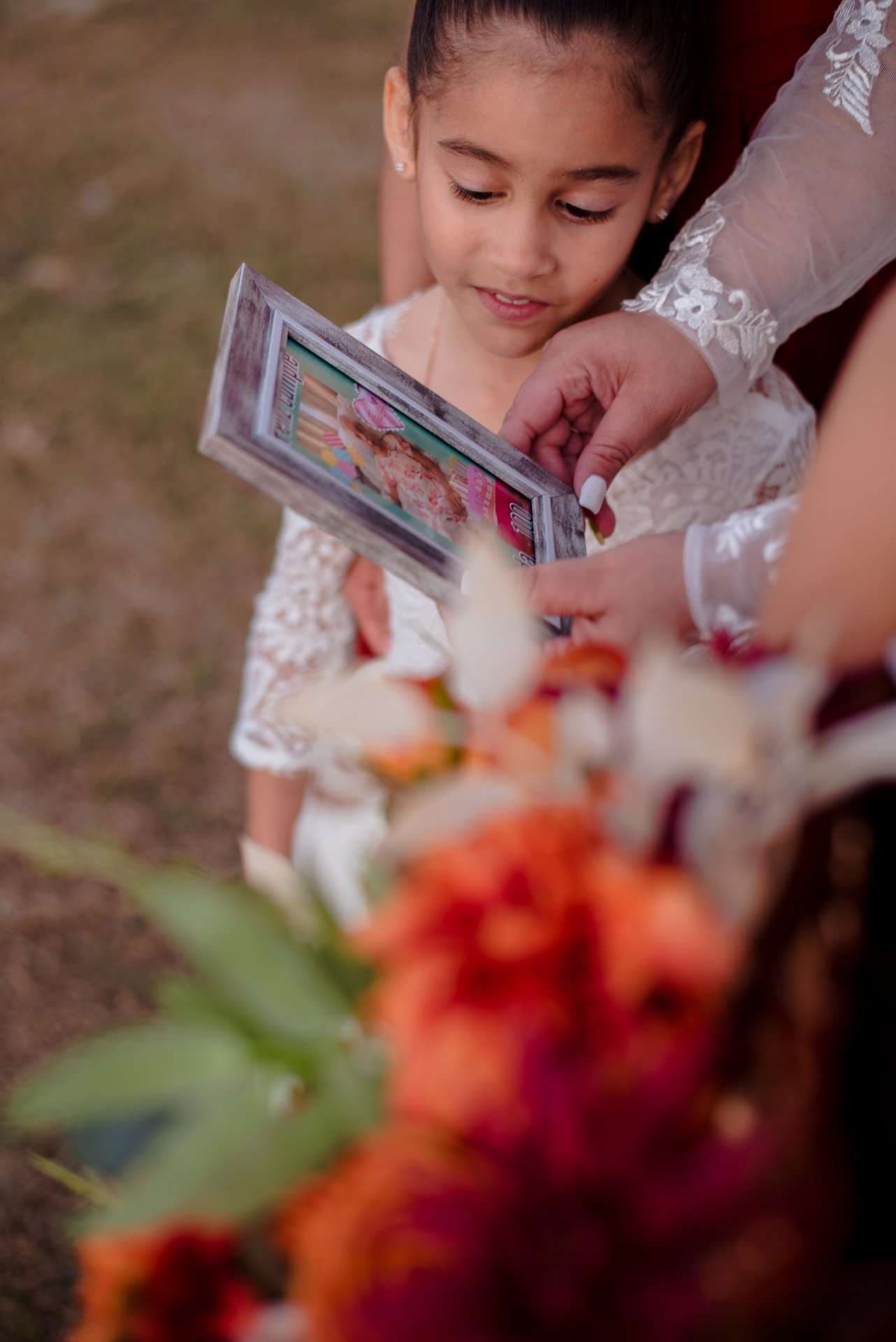 Young girl in a white lace dress looks down at a framed picture held by an adult, with orange and red flowers in front