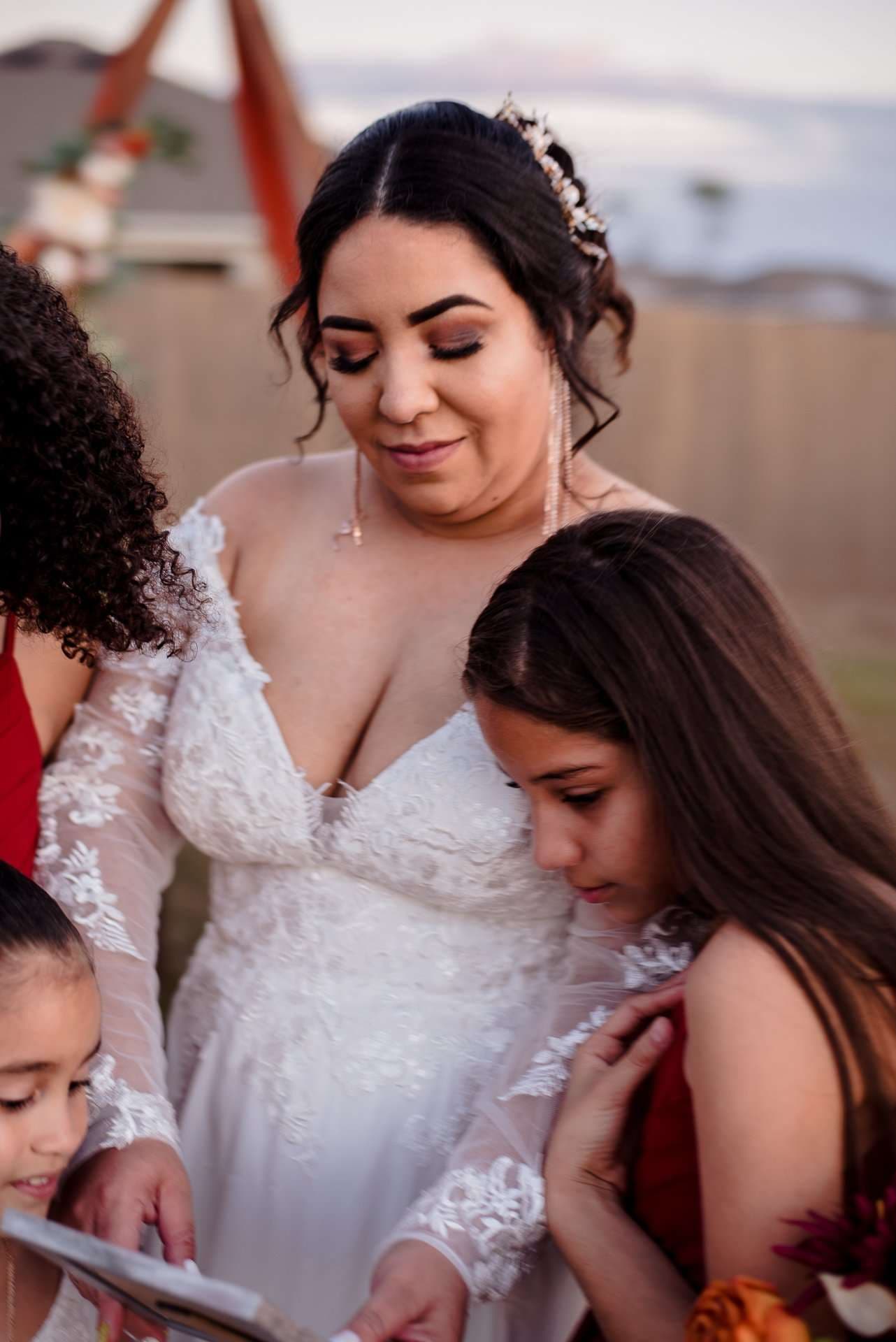 Bride in a white lace gown looks down at a book with two girls in red dresses at an outdoor ceremony