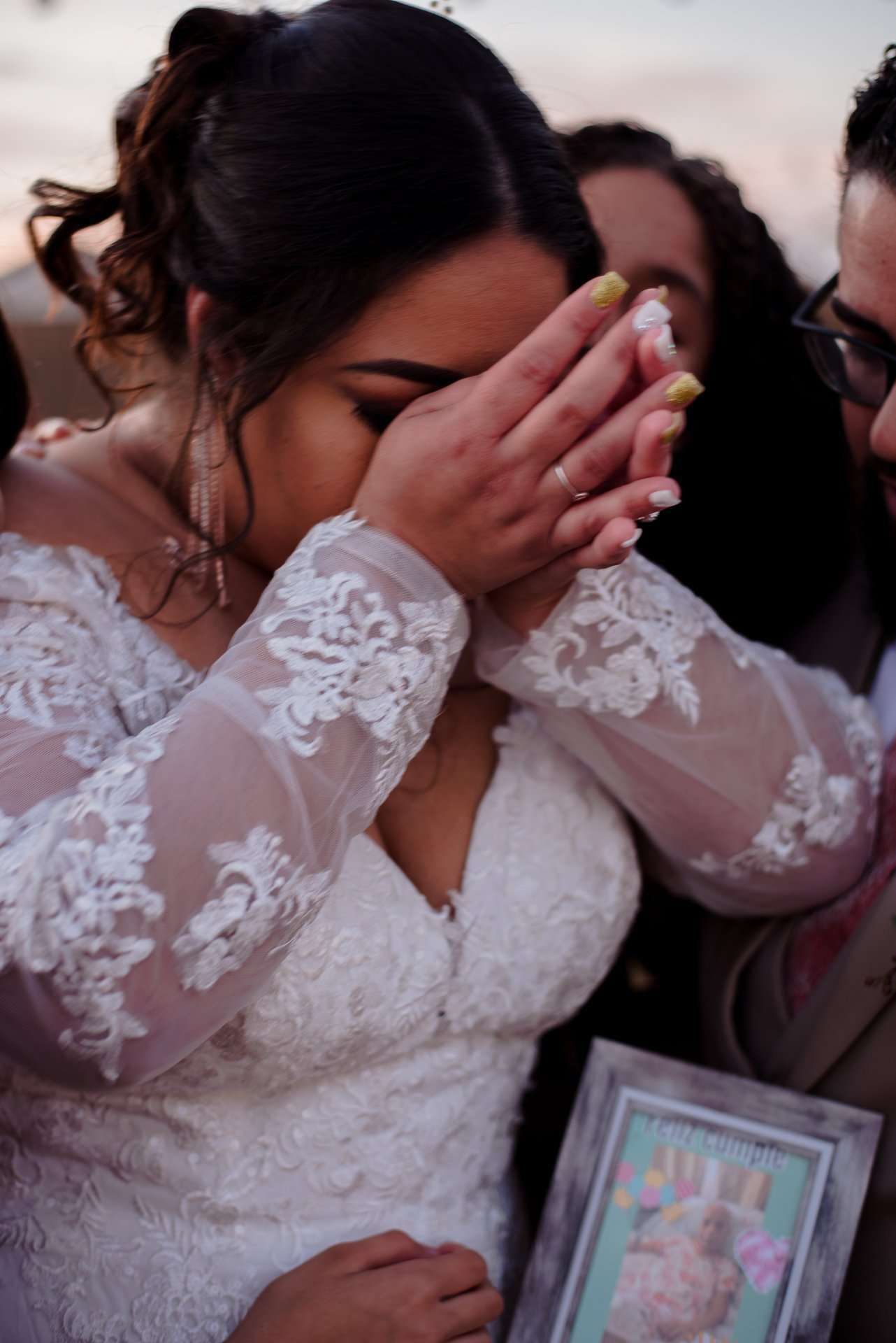 Bride in a white lace gown covers her teary face with both hands while holding a framed picture, surrounded by loved ones