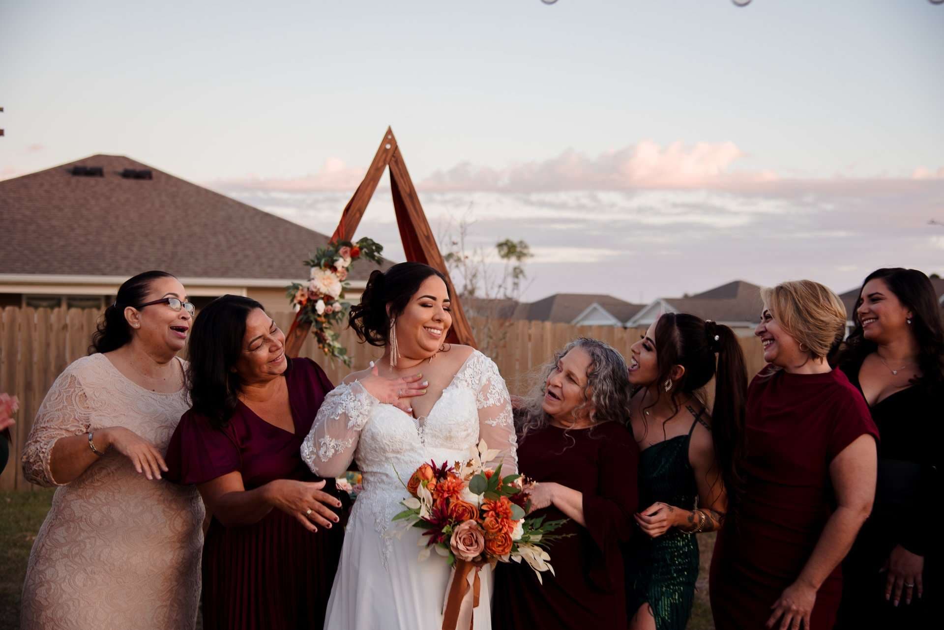 Bride in a white lace gown holding an orange bouquet, laughing with bridesmaids in burgundy beside a wooden triangle arch