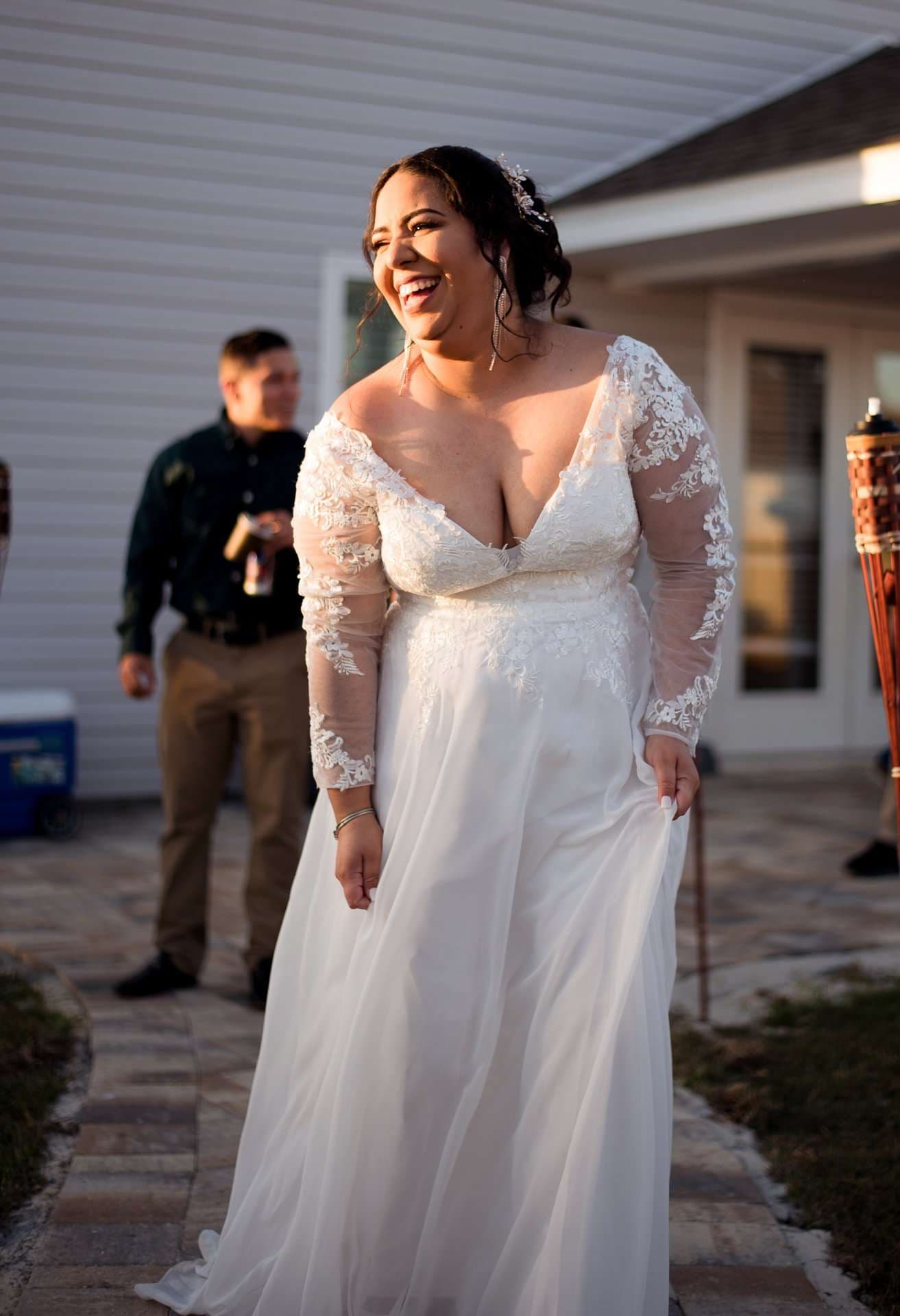Bride in a white lace gown with sheer sleeves laughing outdoors on a brick patio while a guest stands behind her