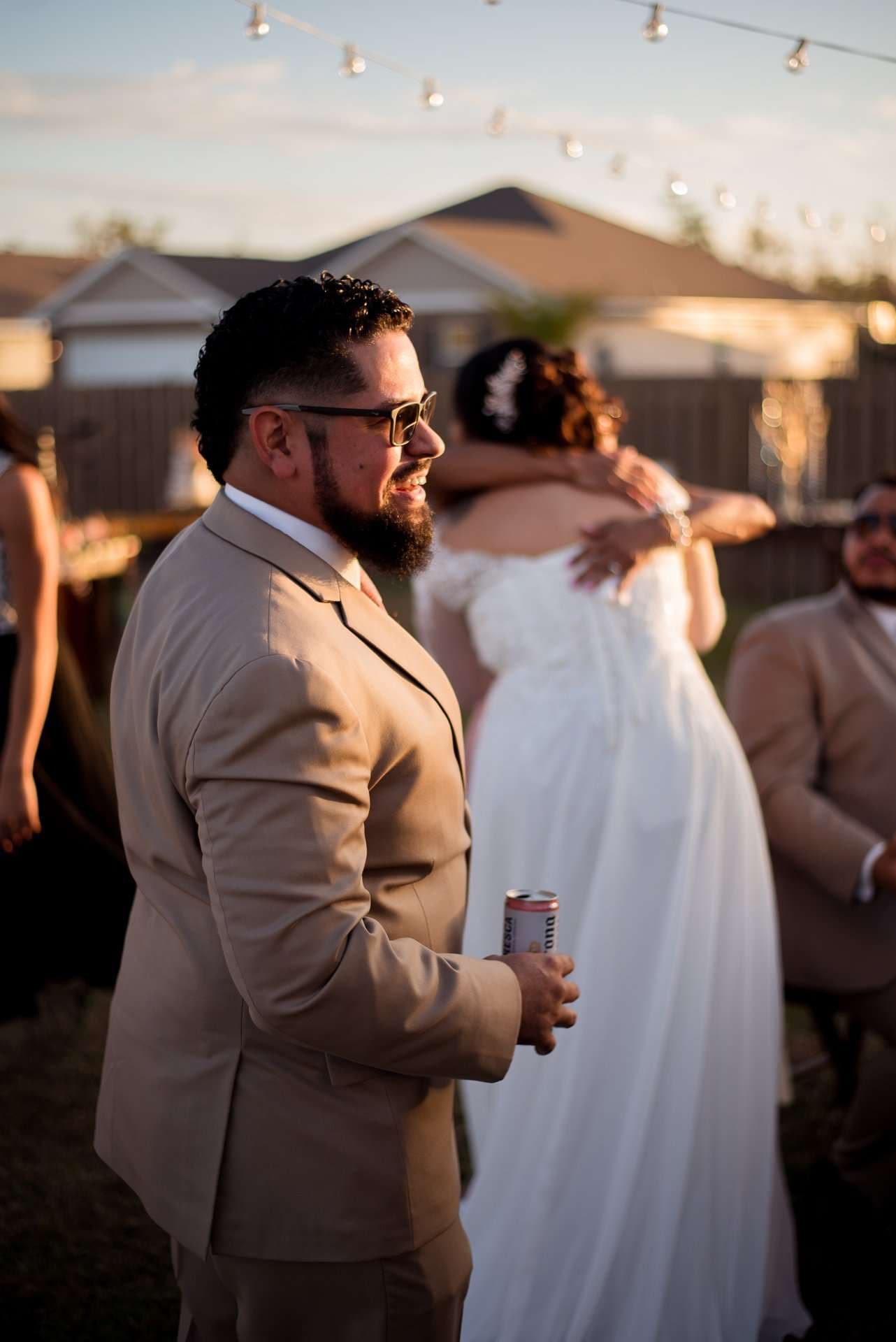 Bearded man in tan suit and sunglasses holds a drink can while a bride in white embraces someone behind him