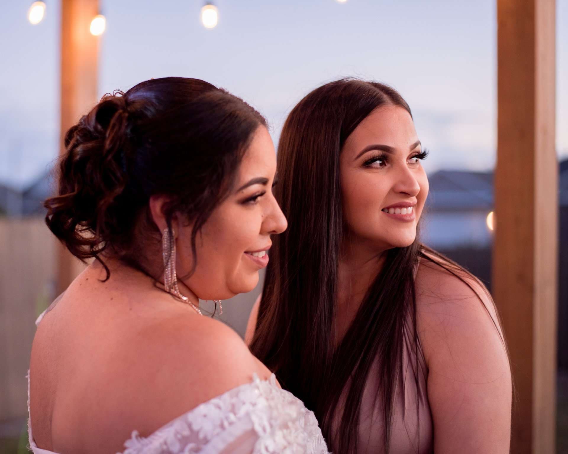 Two smiling women looking off to the side under string lights, one in an off-shoulder white lace gown with an updo