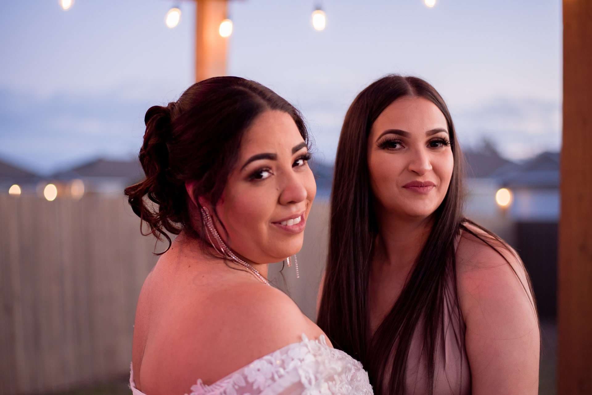Two women posing outdoors at dusk under string lights, one in an off-shoulder white lace gown, the other in a mauve dress