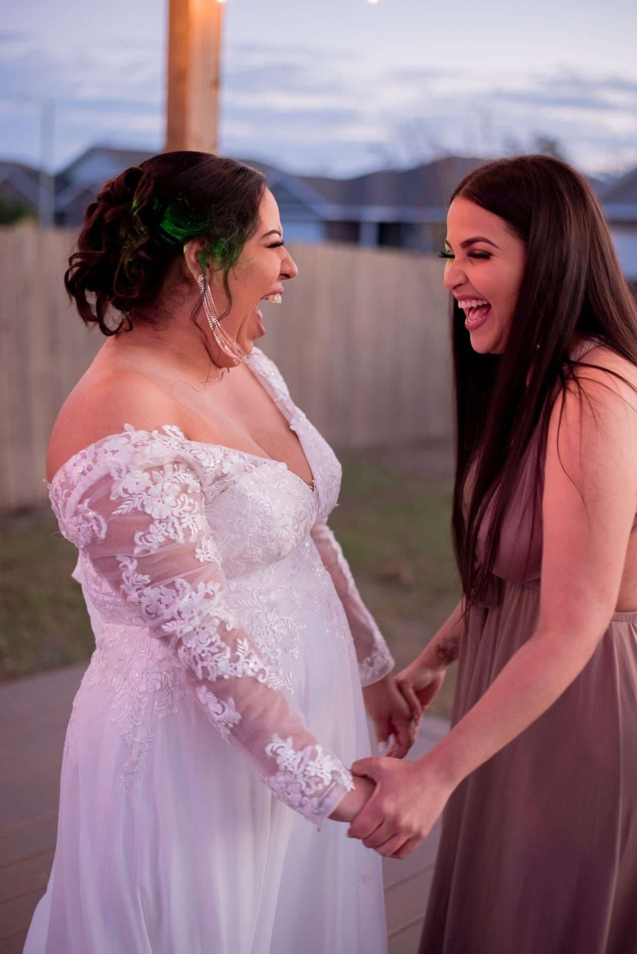 Bride in an off-shoulder white lace gown holding hands and laughing with a woman in a long mauve dress outdoors